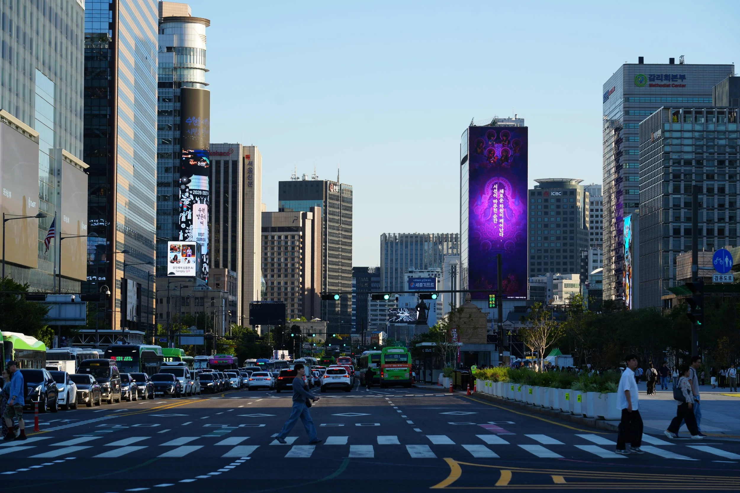 A busy city street with tall buildings, digital billboards, and pedestrians crossing an intersection. Vehicles and green buses are visible on the road, with a blue sky overhead.