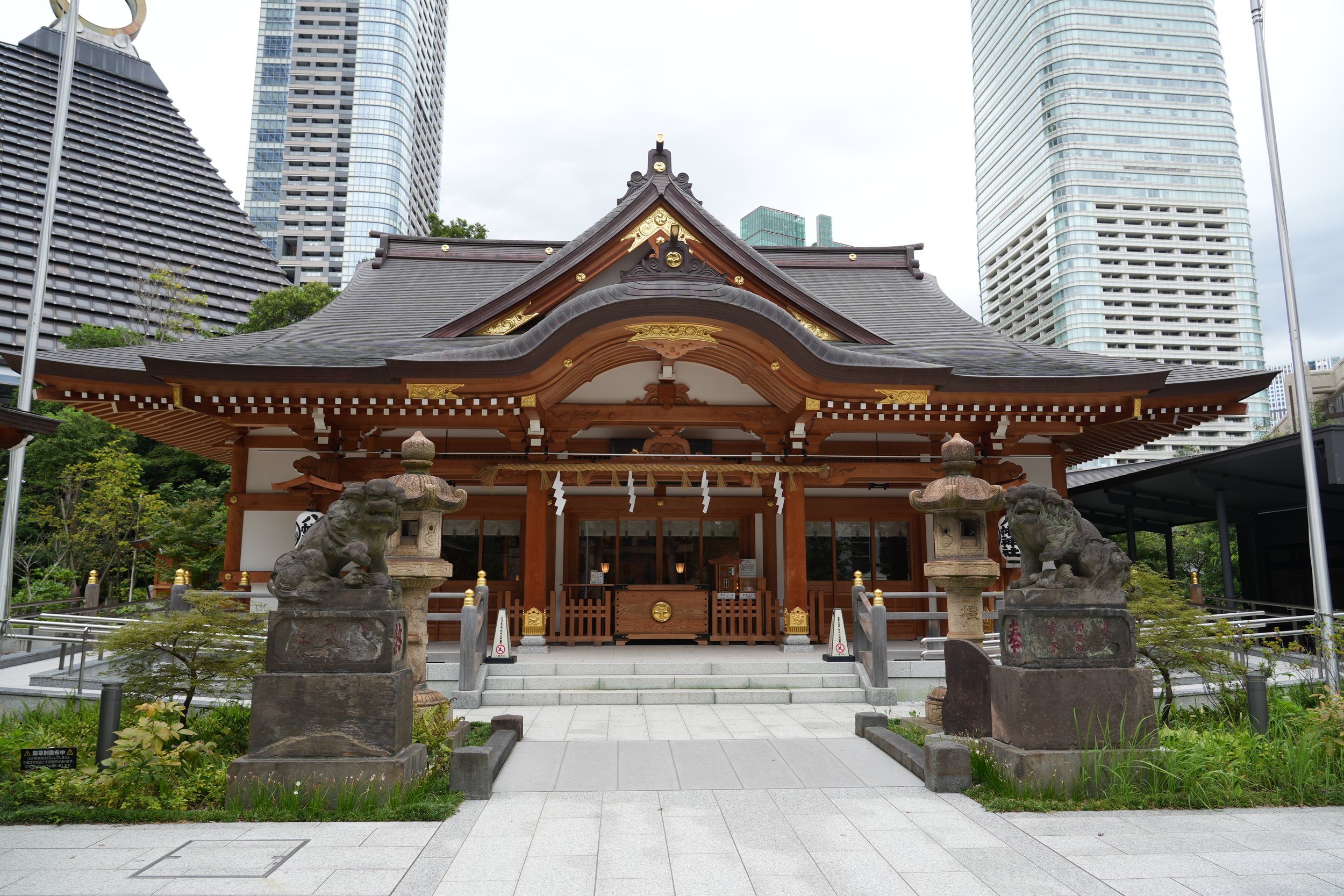 A traditional Japanese shrine in an urban area, with modern skyscrapers in the background, stone lanterns and guardian statues at the entrance.