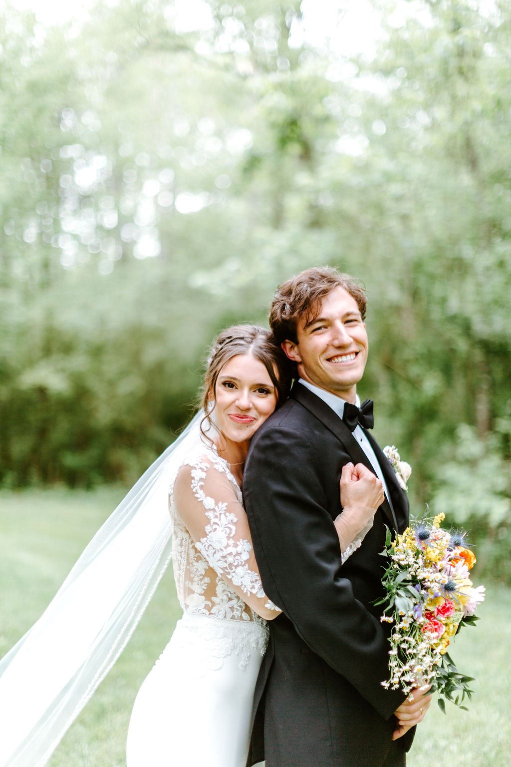 A newlywed couple outdoors on a wedding day, with the bride embracing the groom from behind, holding a bouquet of flowers, both smiling at the camera, surrounded by green trees.