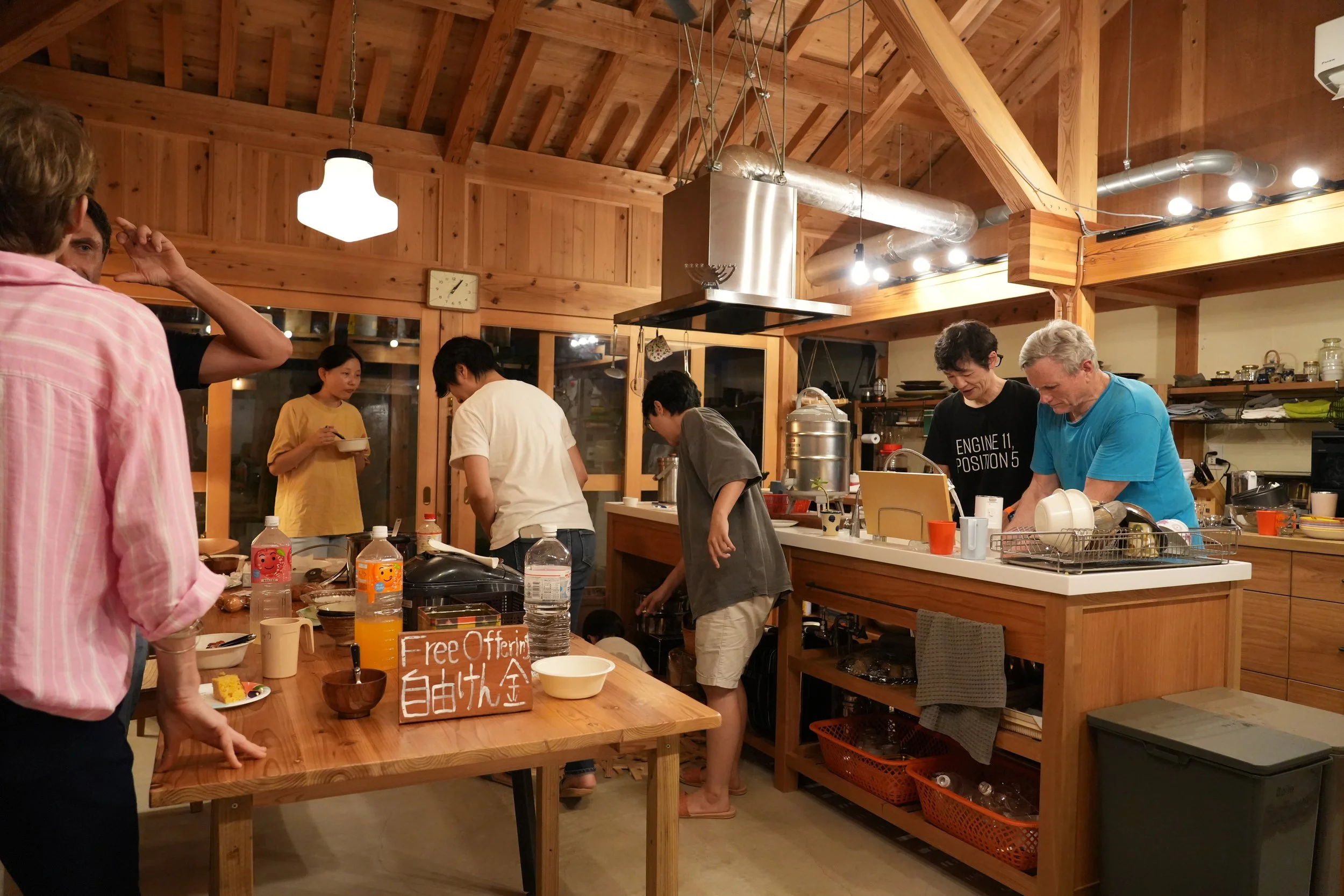 People gathered in a cozy, wooden ceiling kitchen with industrial elements, preparing food and serving drinks, with a sign offering free adult food.