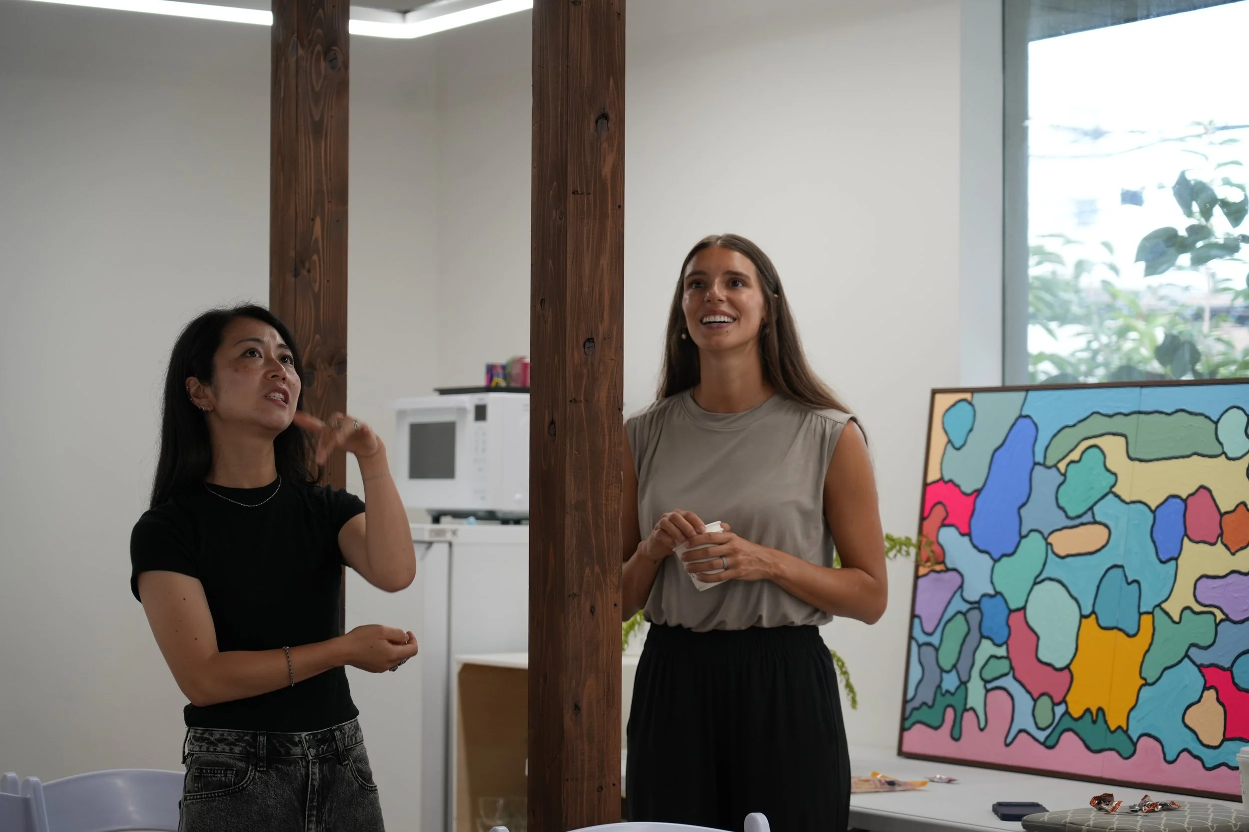 Two women are standing in a room, engaged in conversation. There is a colorful abstract painting on an easel in the background, and a large window with visible greenery outside. A wooden support beam is also visible between the women.