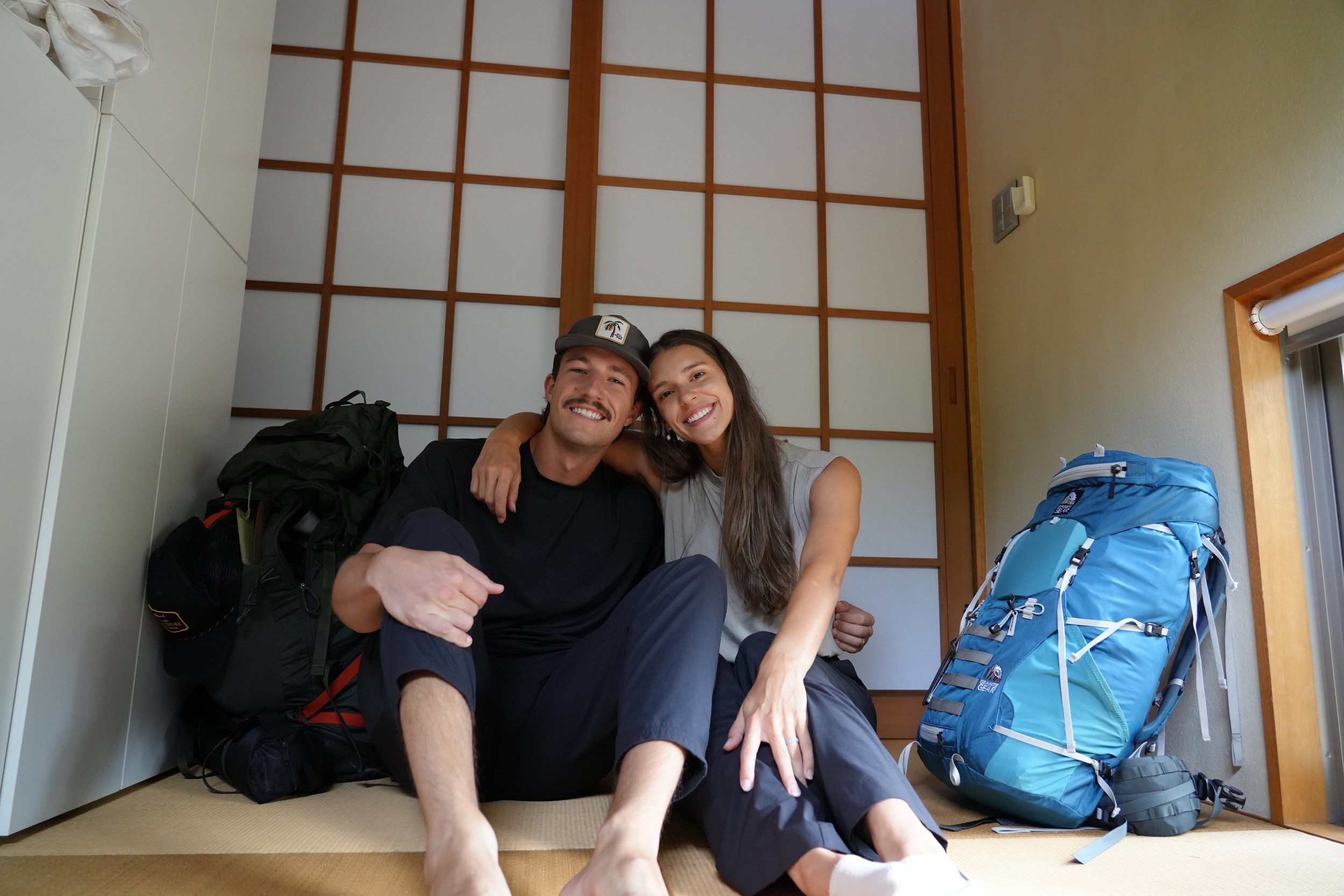 A smiling man and woman sitting inside a room with tatami mats, surrounded by backpacks and gear, with a wooden grid door behind them.
