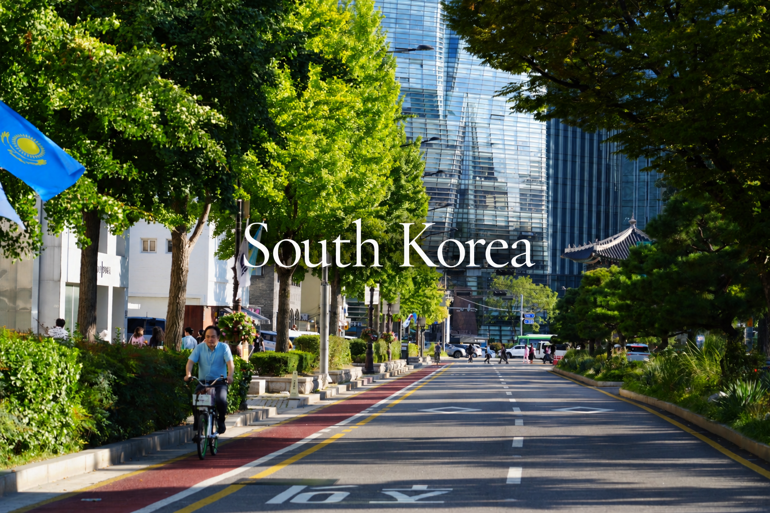 City street in South Korea with modern glass buildings, traditional Korean pavilion, trees, pedestrians, and cyclists.