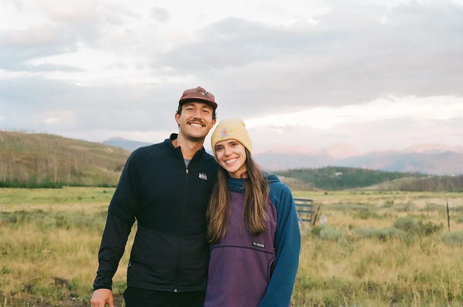 A happy man and woman standing outdoors in a grassy field with hills and cloudy sky in the background, smiling at the camera.