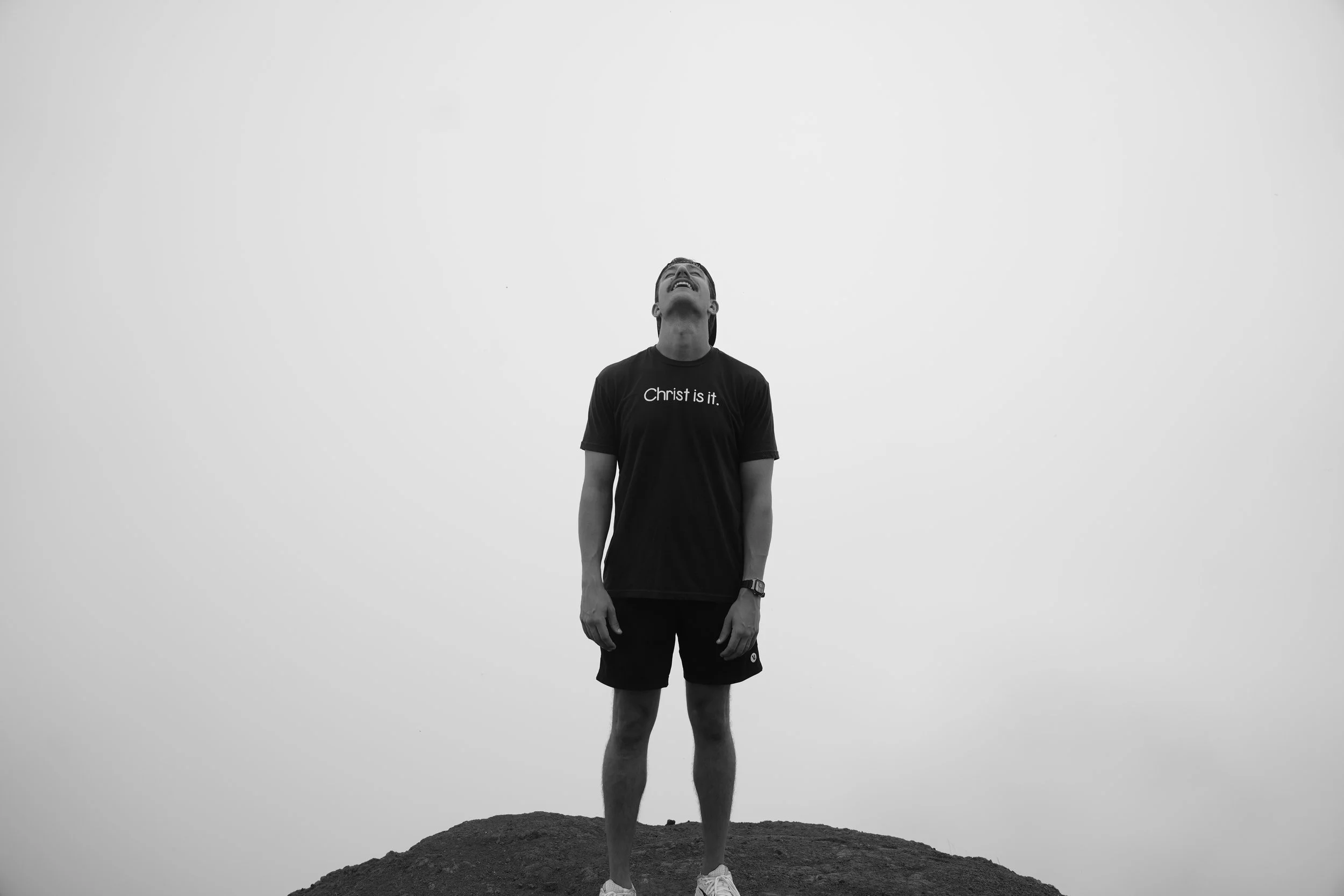A young man standing on a mound of dirt or gravel, looking upwards with a joyful expression, wearing a black T-shirt that says 'Christ is it.' and black shorts.