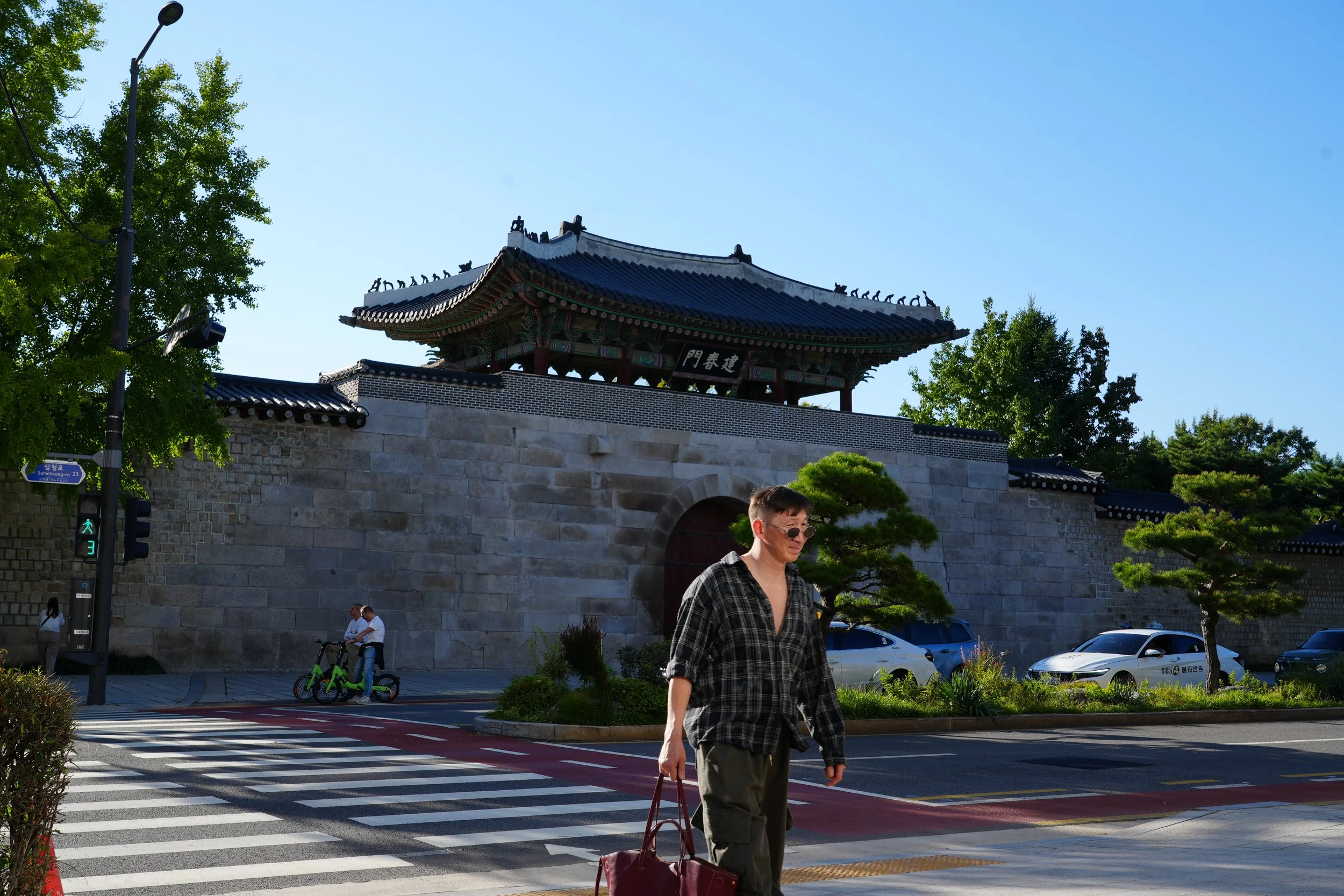 A traditional East Asian building with ornate roof and decorative elements, situated above a stone wall, with green trees around it. There are a few people walking on the street, including a woman in the foreground with sunglasses and a plaid shirt, 