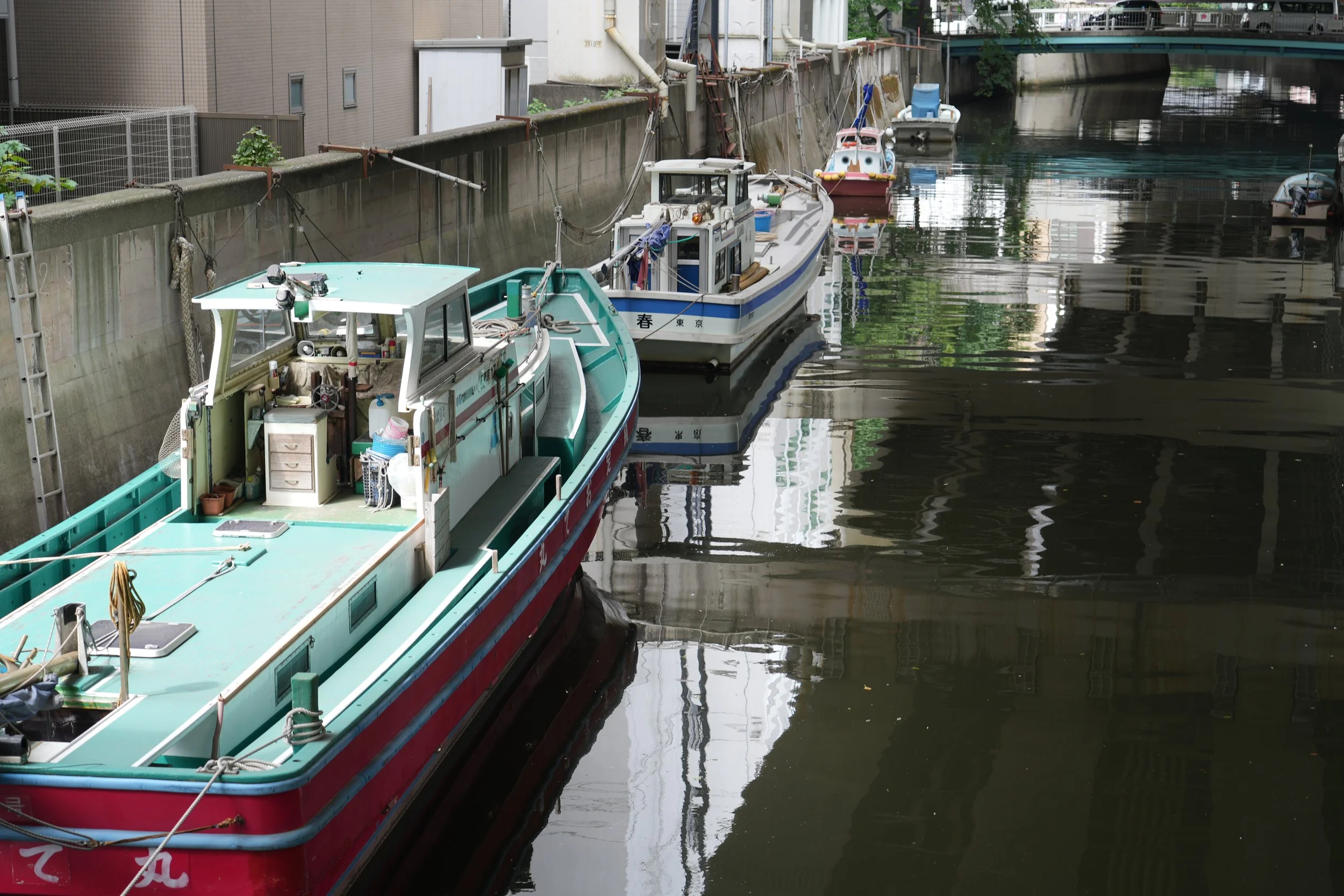Several boats docked along a narrow, calm canal with dark water, surrounded by urban buildings and a bridge overhead.