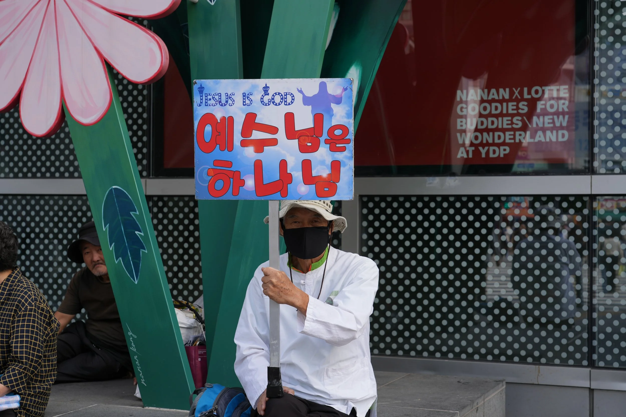 A person wearing a white shirt, black face mask, and hat is sitting on a bench holding a sign that reads 'Jesus is God' with Korean characters below. There are other people sitting nearby and colorful decorations around.