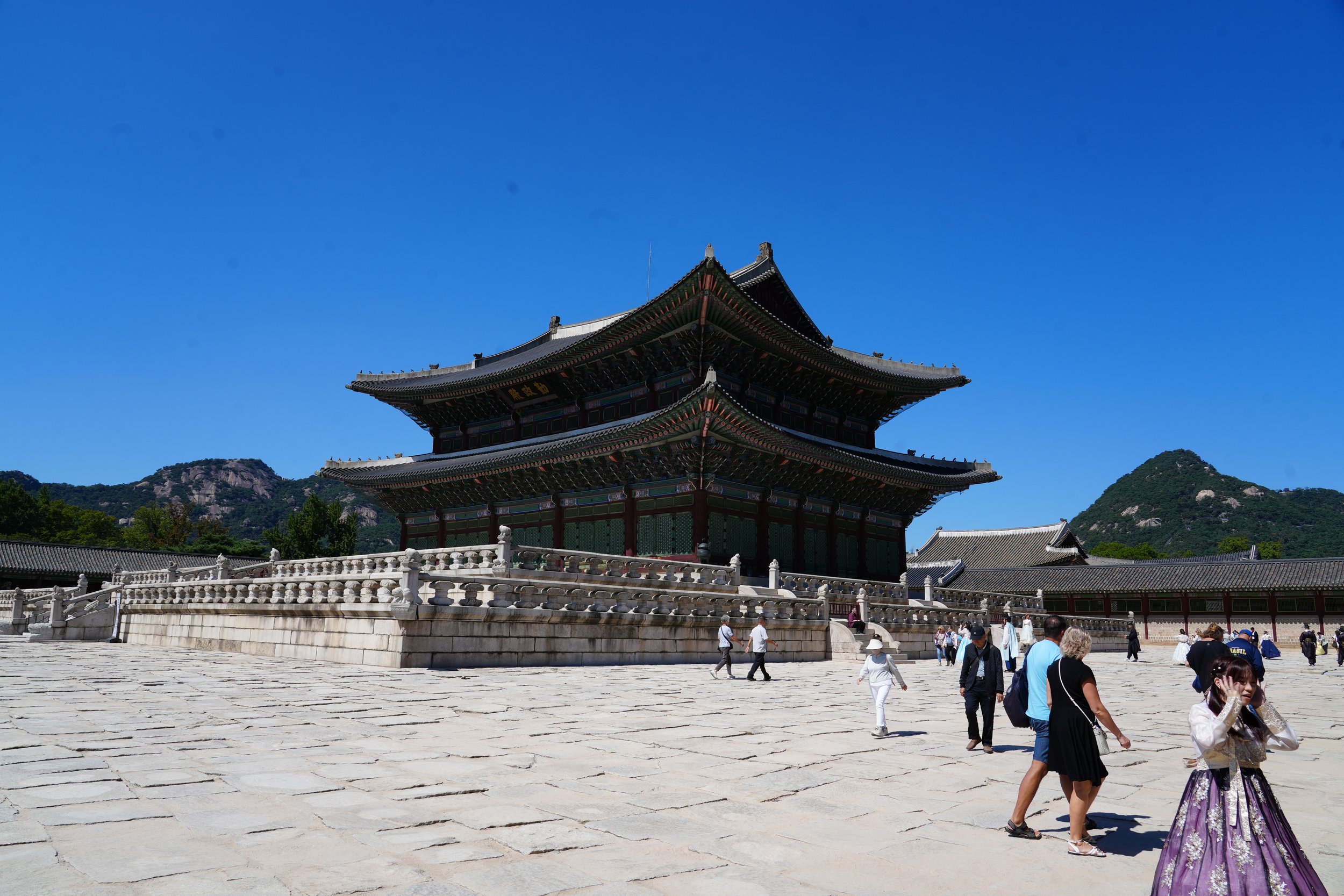 Tourists walking in front of a traditional Asian temple with a multi-tiered, pagoda-style roof, set against mountainous landscape under clear blue sky.