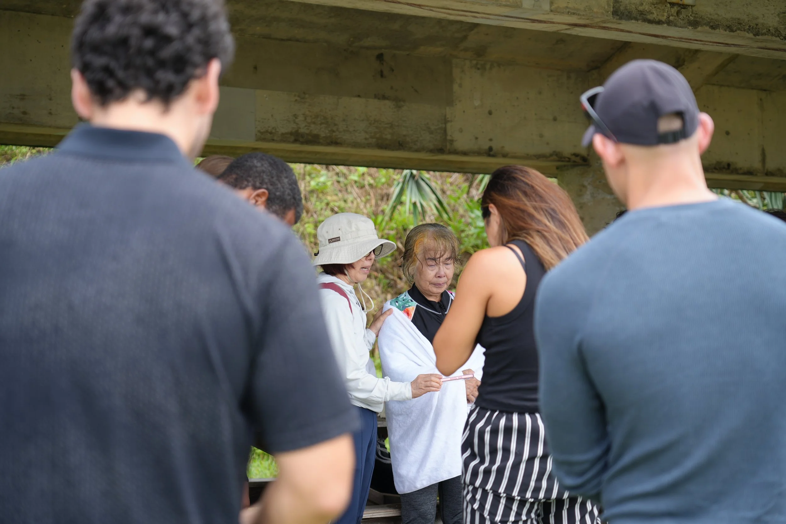 A group of people gathered under a bridge, with a few women looking at a phone and others listening, surrounded by greenery.