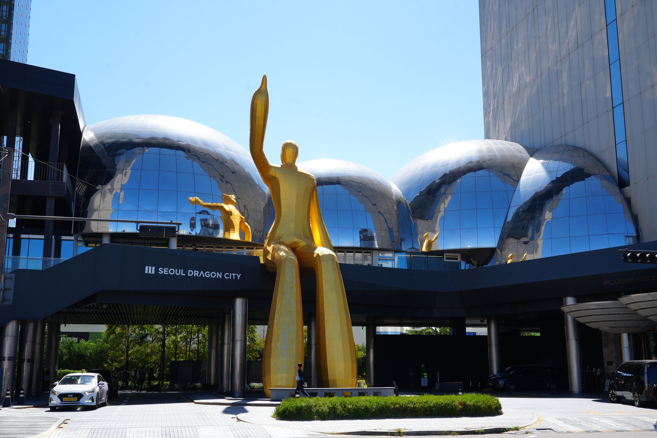 Golden sculpture of a person with raised arm in front of a modern building with reflective silver domes in Seoul, South Korea.