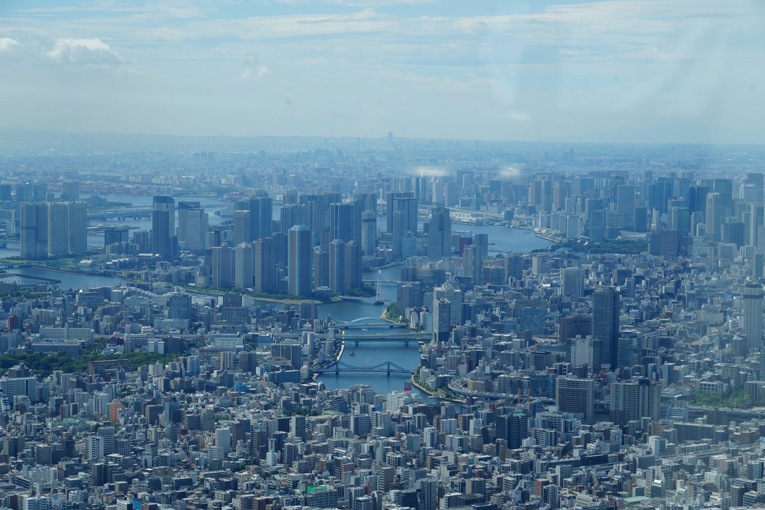 Aerial view of a city skyline with numerous high-rise buildings, a river running through the city, and a bridge crossing the river, under a partly cloudy sky.