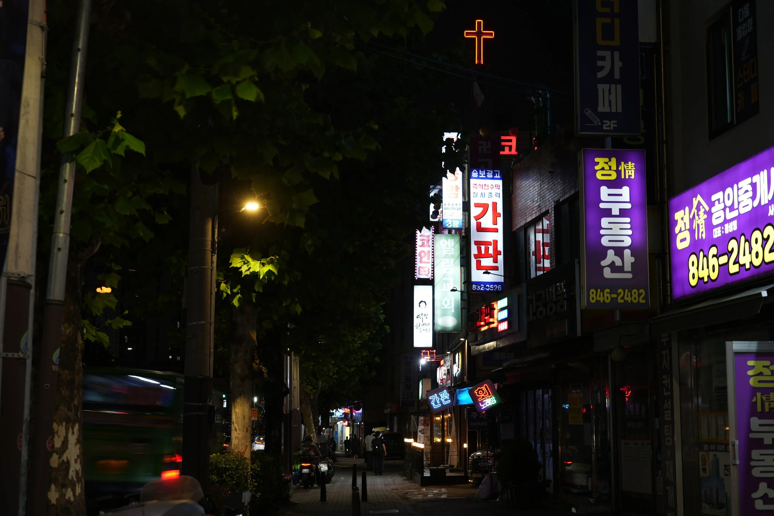 Night street scene with illuminated neon and electronic signs in various languages, some in Korean, hanging on buildings. Trees line the sidewalk, and people walk along the street. There are also cars and a bus in motion, creating light streaks.