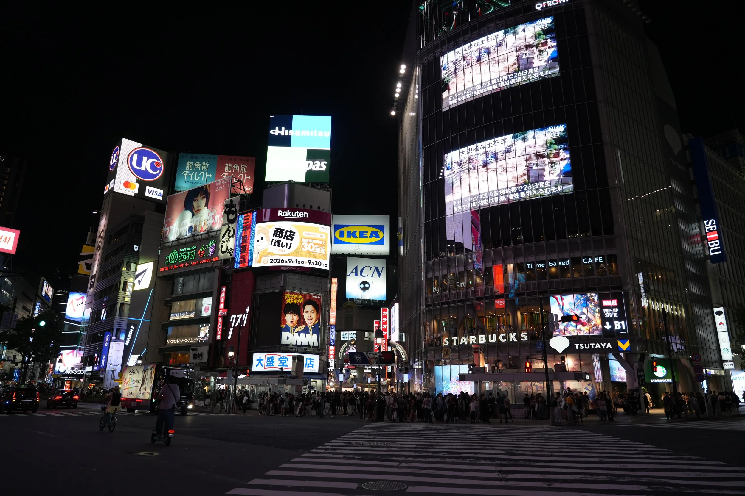 Nighttime city scene with tall buildings covered in bright digital billboards and signs, pedestrians crossing the street, and cars visible in the traffic.