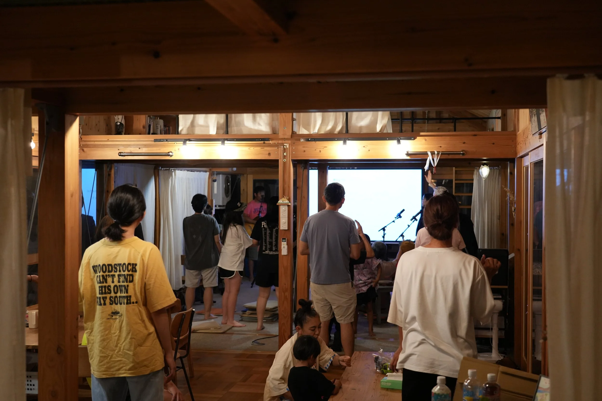 People gathered in a cozy wooden room, some standing and some sitting, watching a presentation or performance on a large screen, with microphones set up in front of the screen.