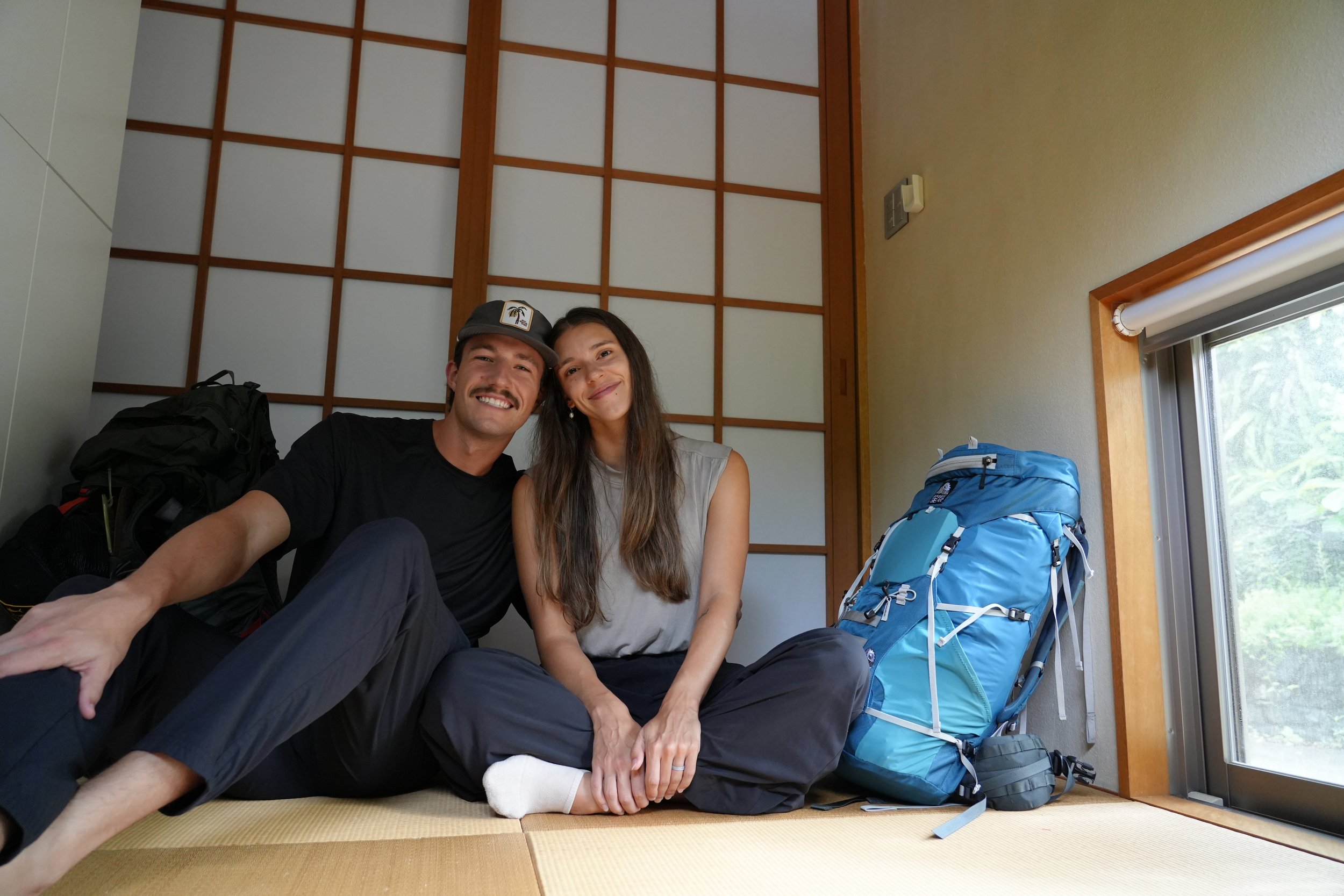 A smiling man and woman sitting on the floor in a room with traditional Japanese shoji screens, surrounded by hiking backpacks and gear.