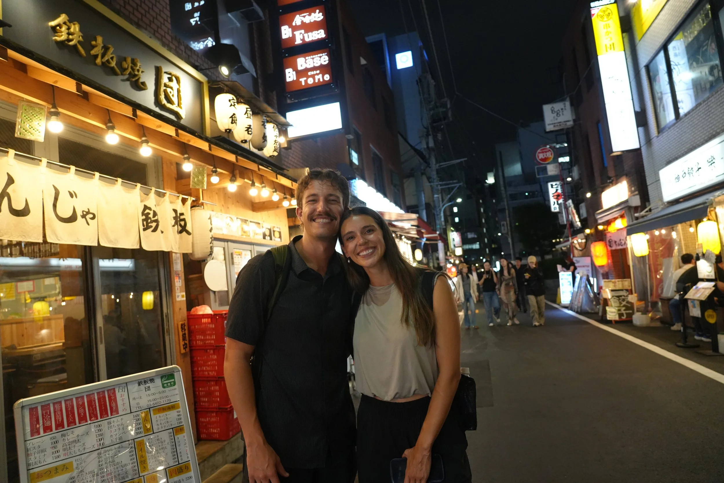 A smiling man and woman standing close together on a busy street at night in Japan. The background shows illuminated signs and shop fronts with Japanese characters, lanterns, and people walking.