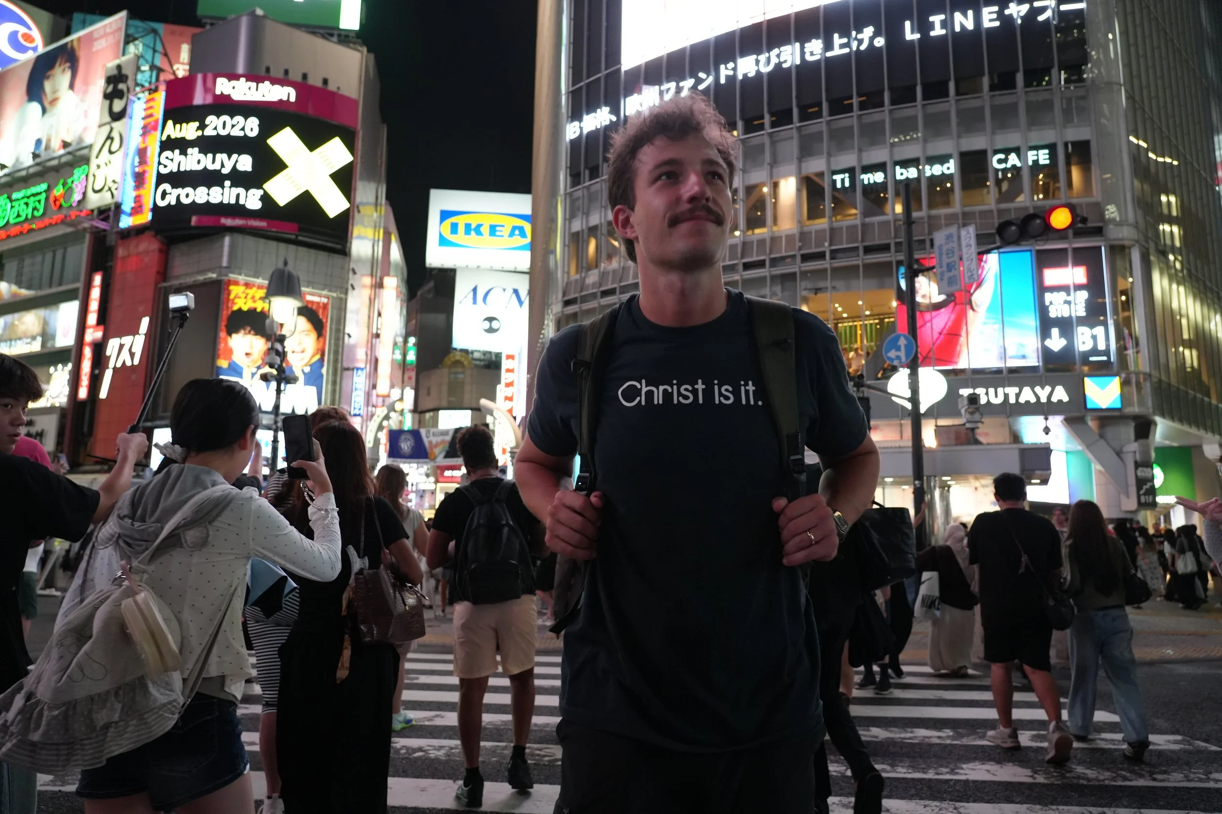 A young man with a mustache and backpack standing at a busy, illuminated intersection in Shibuya, Tokyo, at night, surrounded by other pedestrians and bright electronic billboards.