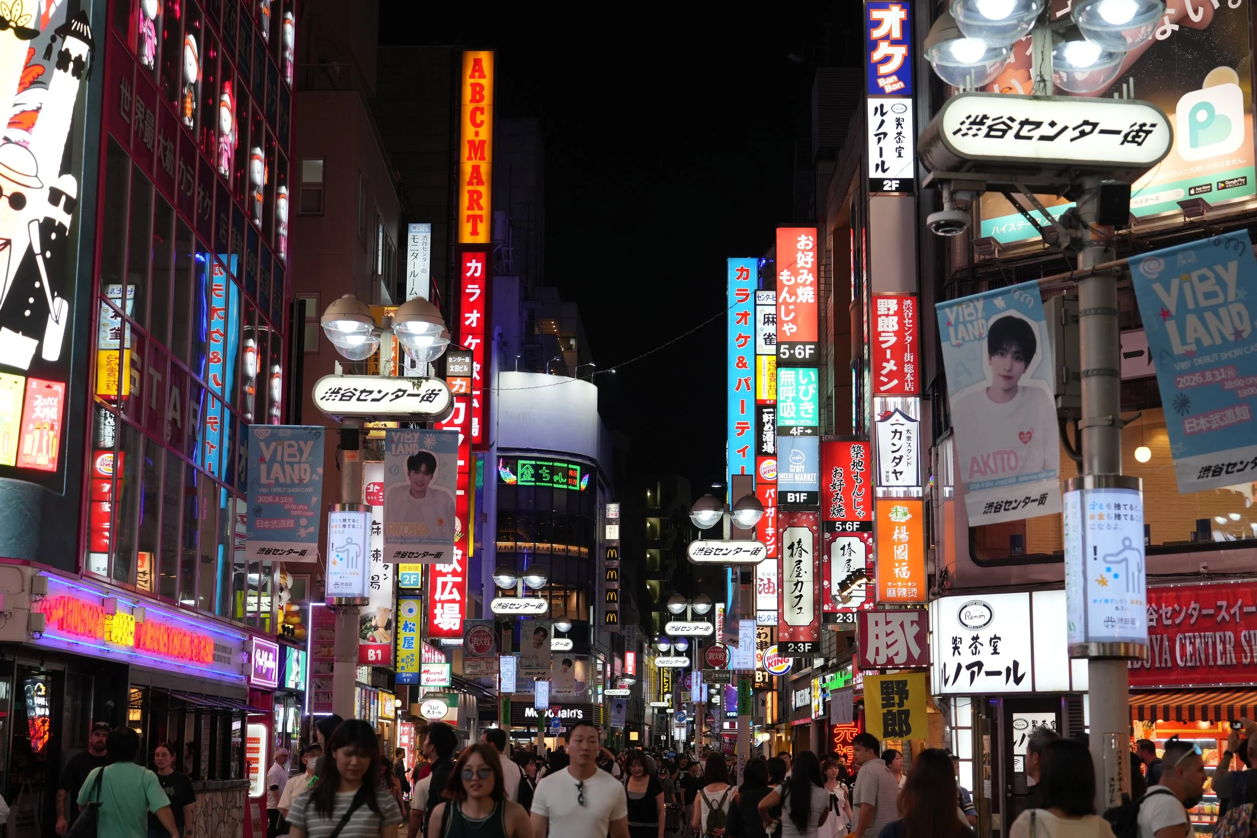 Nighttime street scene in a busy city with colorful illuminated signs in Japanese and English, crowded with pedestrians.