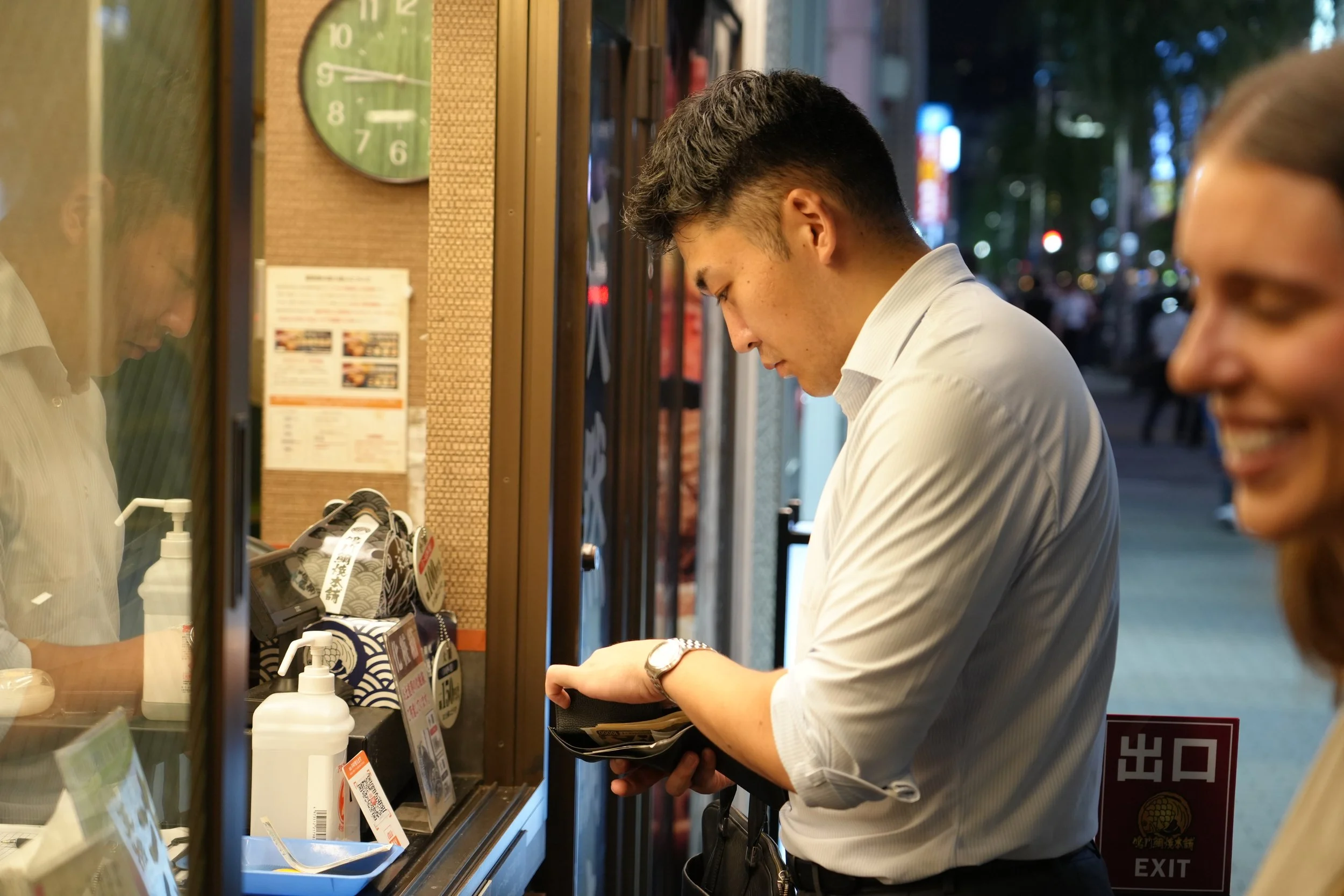 A young man with dark hair wearing a light-colored dress shirt and a wristwatch stands at a counter inside a restaurant or food establishment, looking down at his wallet. To his right is a young woman with glasses, smiling and looking at him. The sce