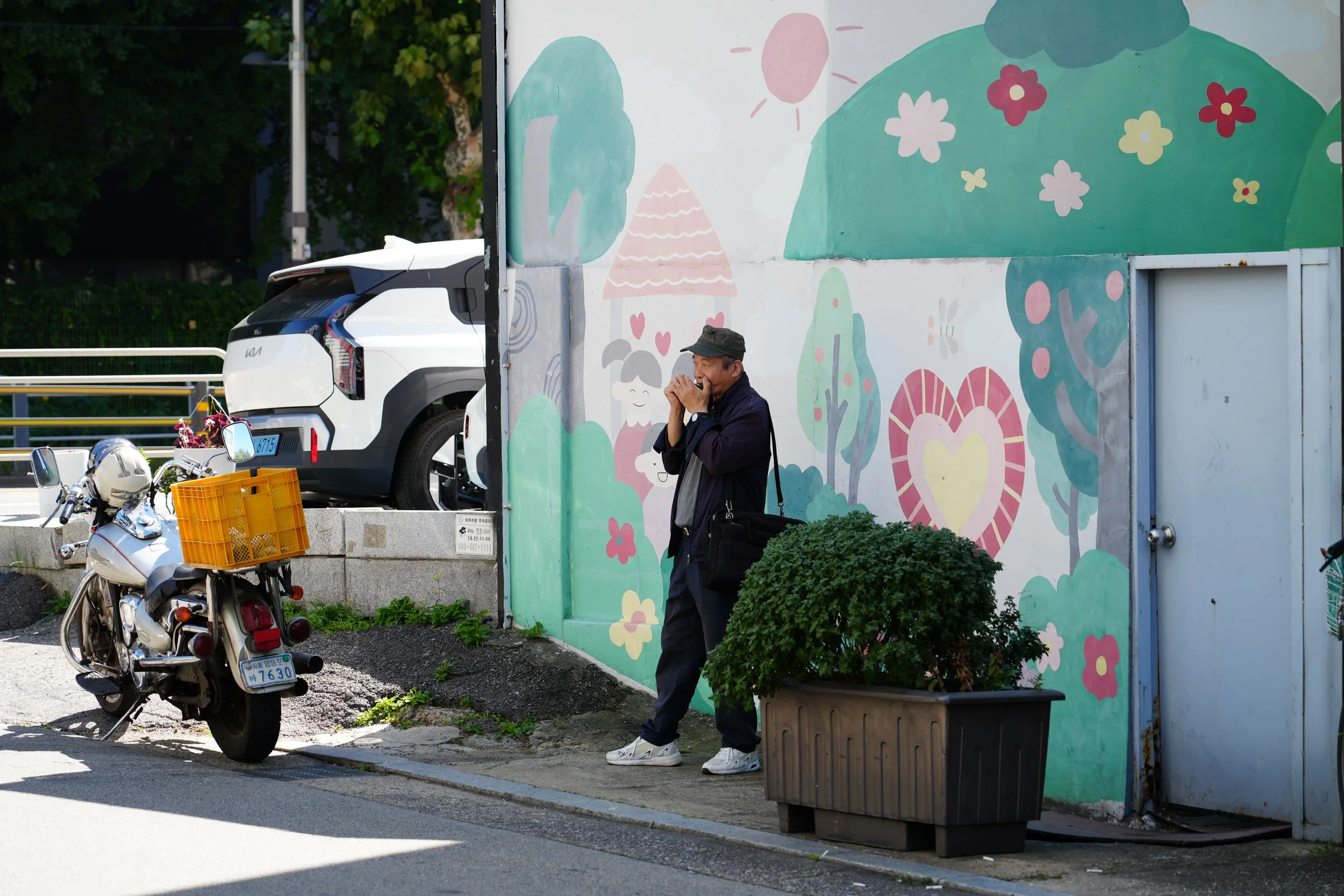 A man standing on a sidewalk near a large painted mural on a wall, with a motorcycle and a white car parked nearby. The mural features colorful, whimsical illustrations of trees, a house, a bunny, and flowers. The man is wearing a cap, dark jacket, a