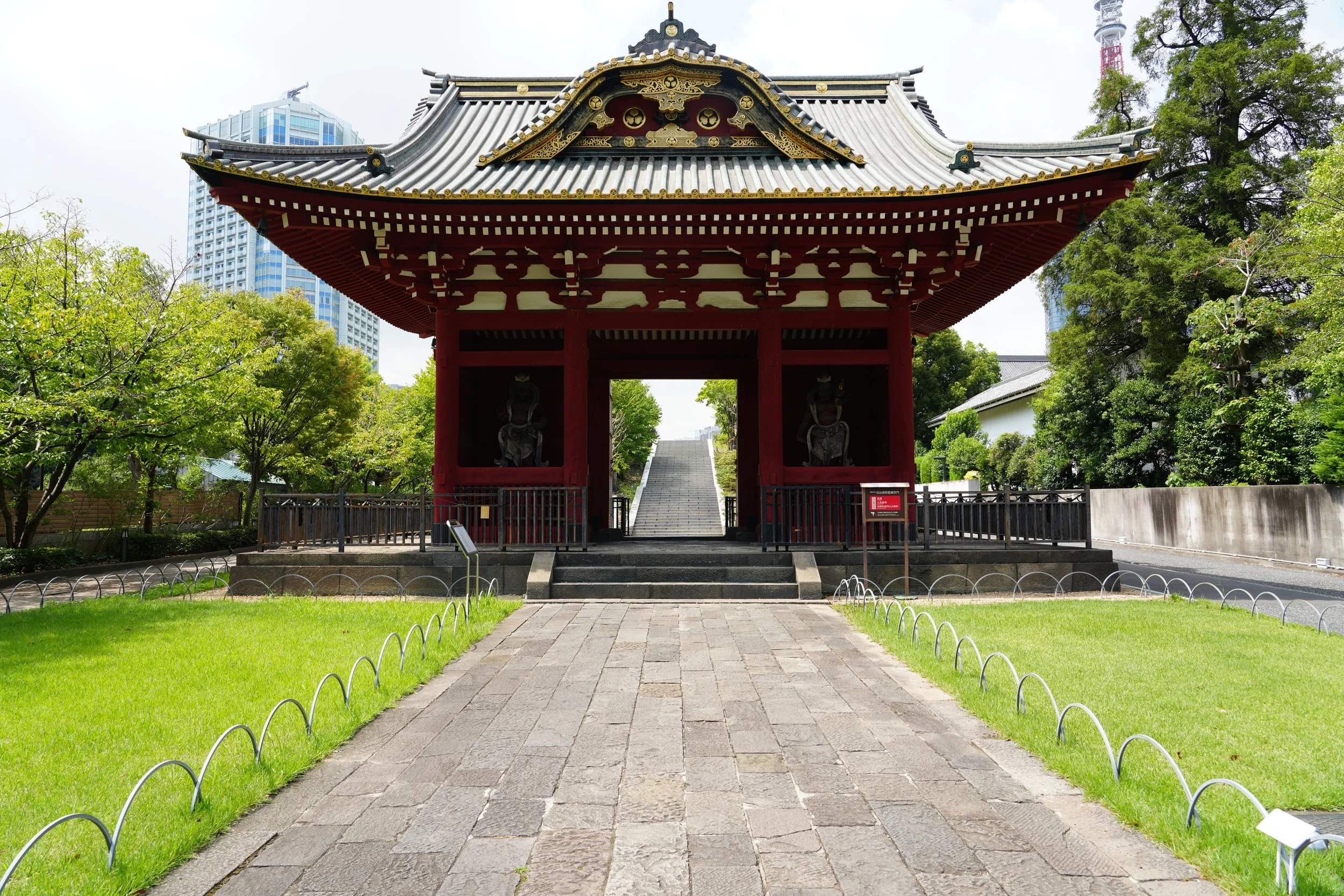 Traditional Japanese temple gate with a curved roof, red pillars, and decorative gold accents, surrounded by green trees and modern buildings in the background.