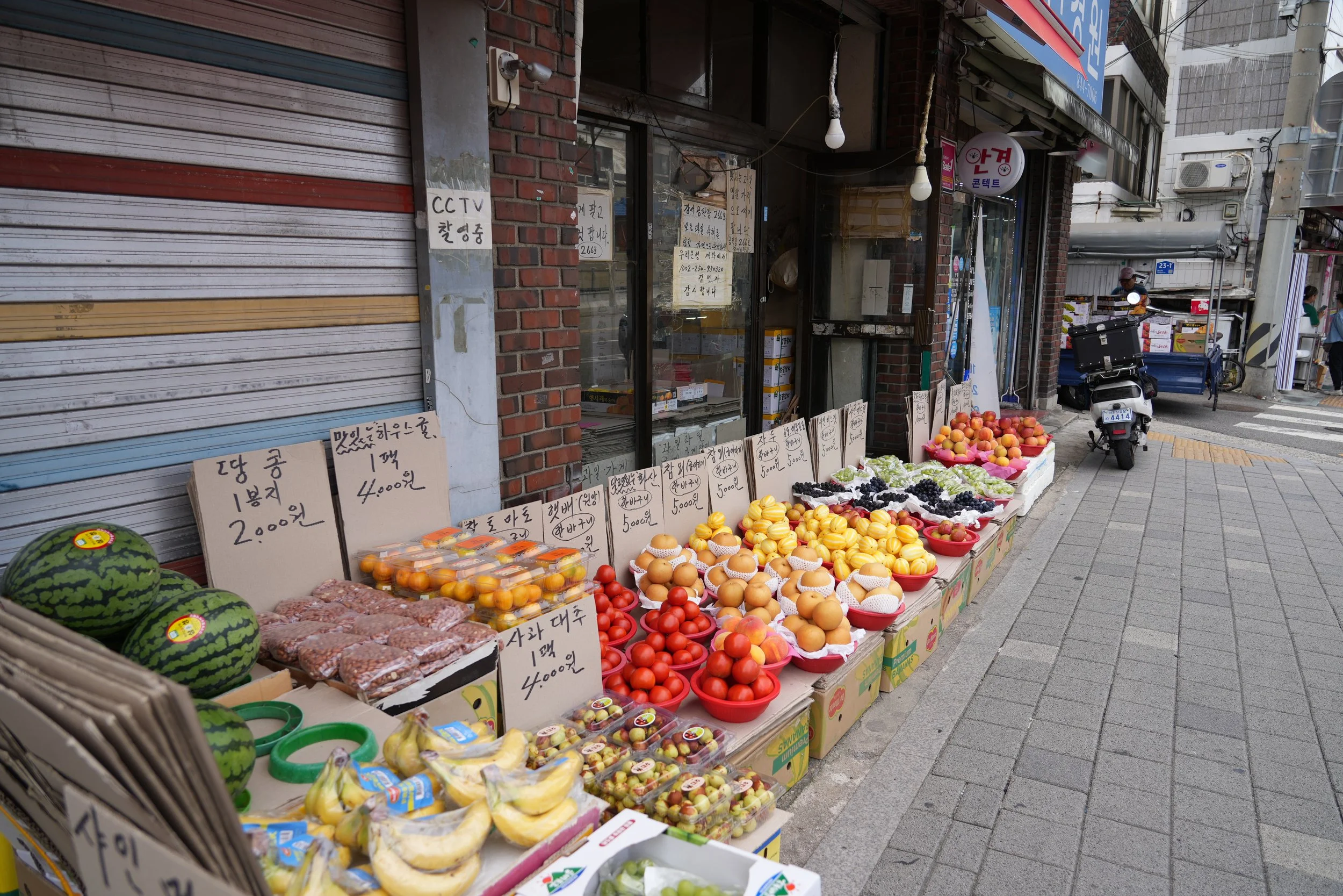 Street market stall selling various fruits, including watermelons, peaches, tomatoes, and grapes, with handwritten signs in Korean and a closed shutter on the left side, in an urban setting.