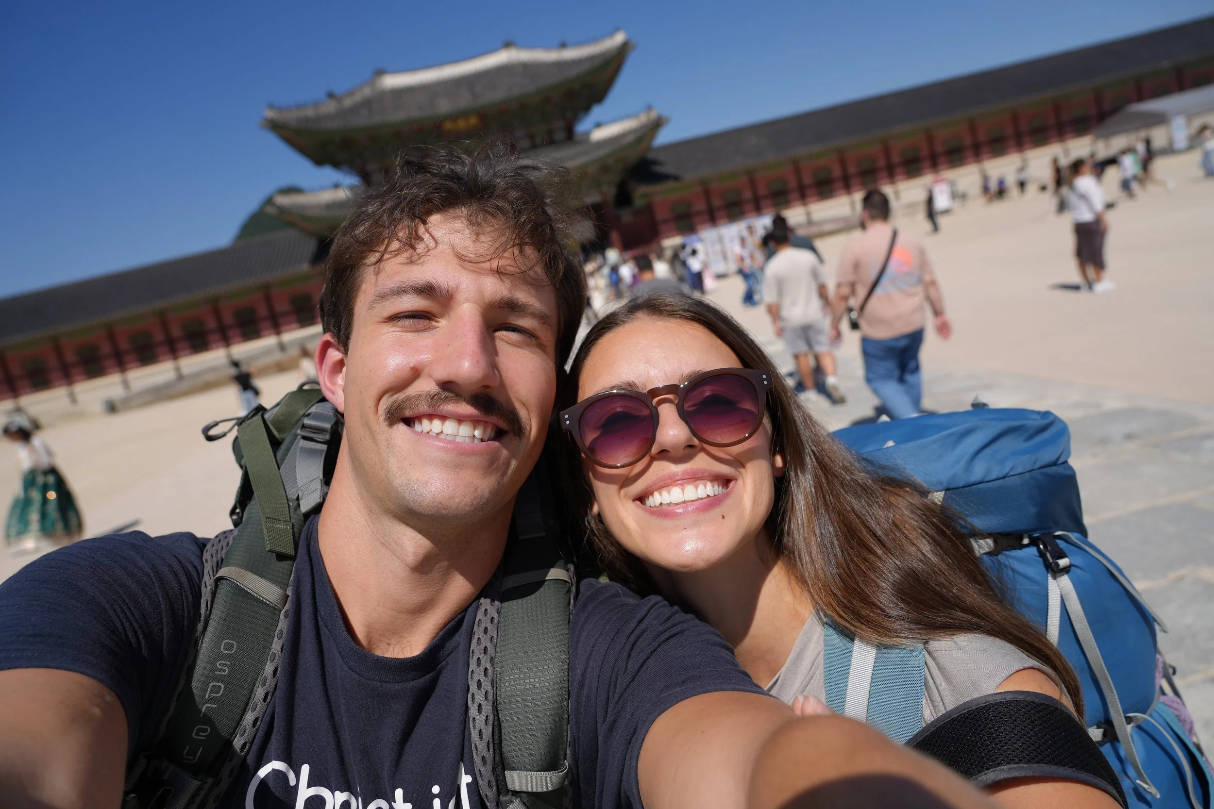 Smiling young man and woman taking a selfie in front of a traditional Asian temple on a sunny day, with other tourists in the background.
