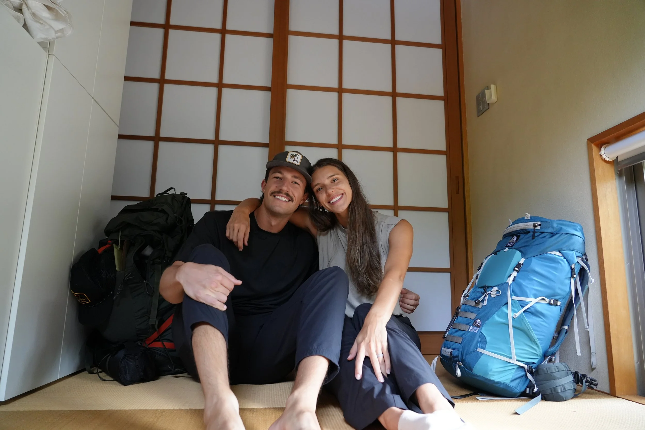A man and woman sitting on the floor inside a room, smiling at the camera, with backpacks and a blue hiking backpack near them. The man is wearing a cap and has a mustache, while the woman has long dark hair.