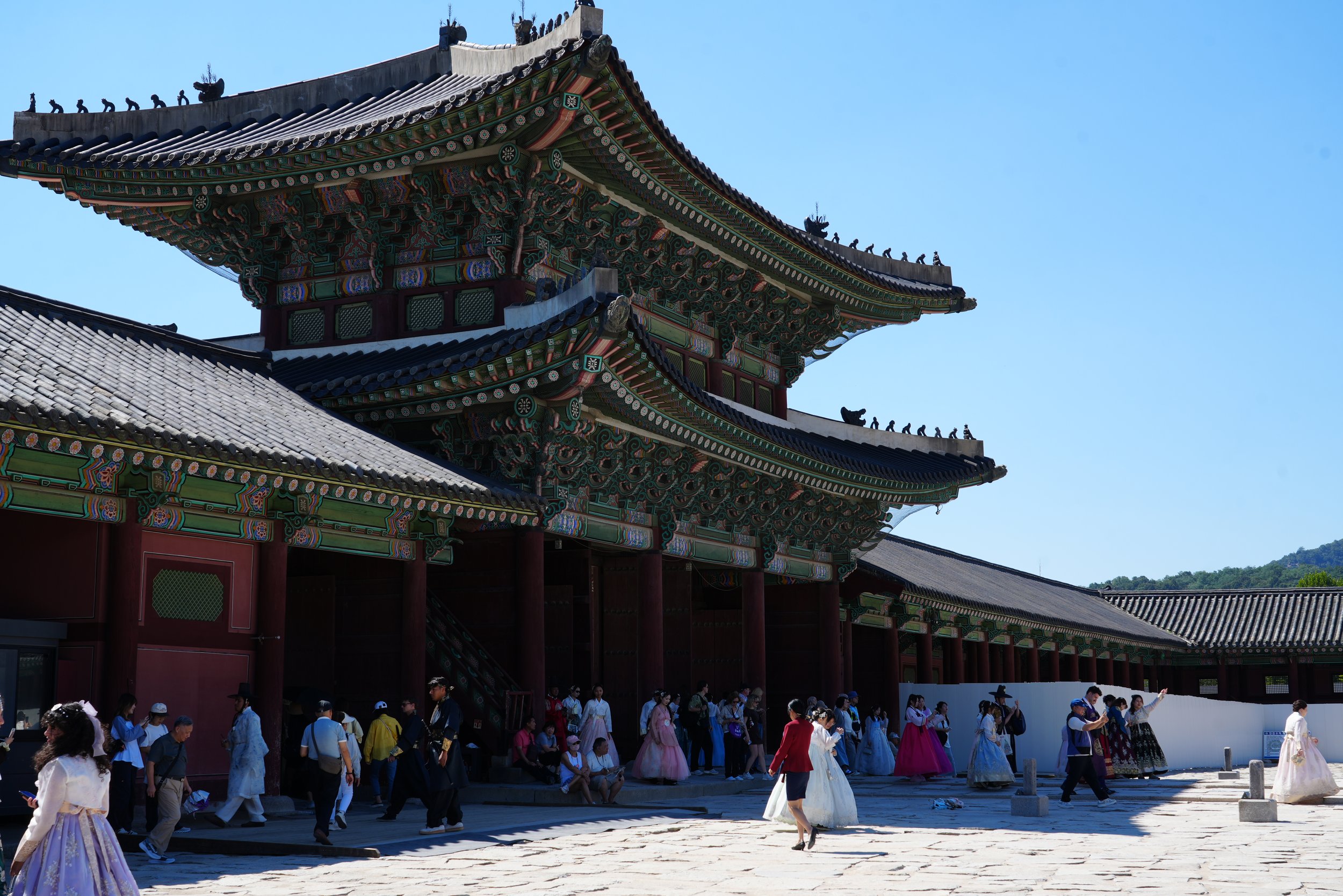 Crowd of people, some in traditional Korean attire, gather outside a colorful, historic Korean palace with ornate multi-tiered roofs and decorative details.