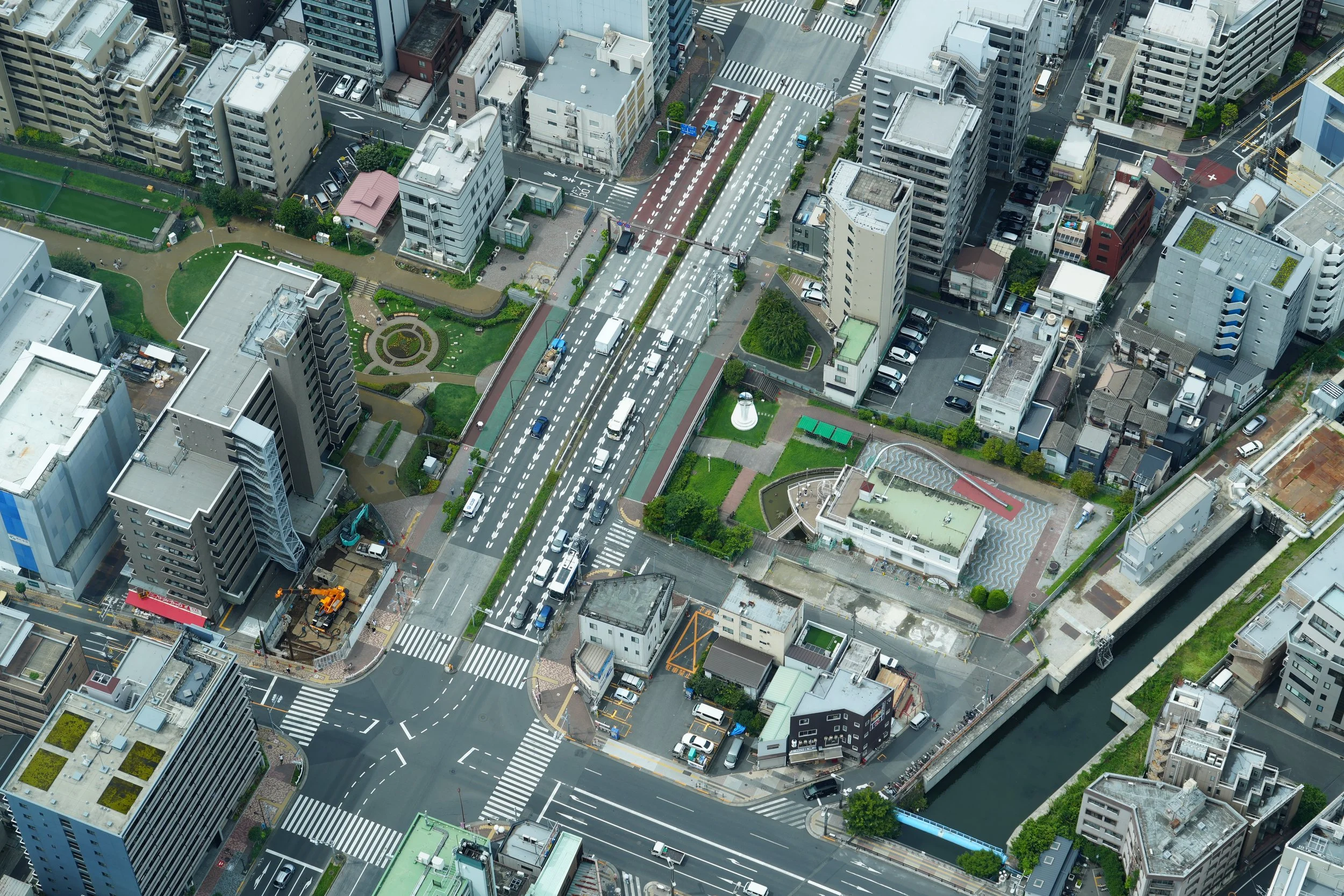Aerial view of a city with tall buildings, streets, and intersections. Vehicles are traveling on the roads, and there are crosswalks and traffic signals visible. Some green spaces and small parks are also present amidst the high-rise structures.