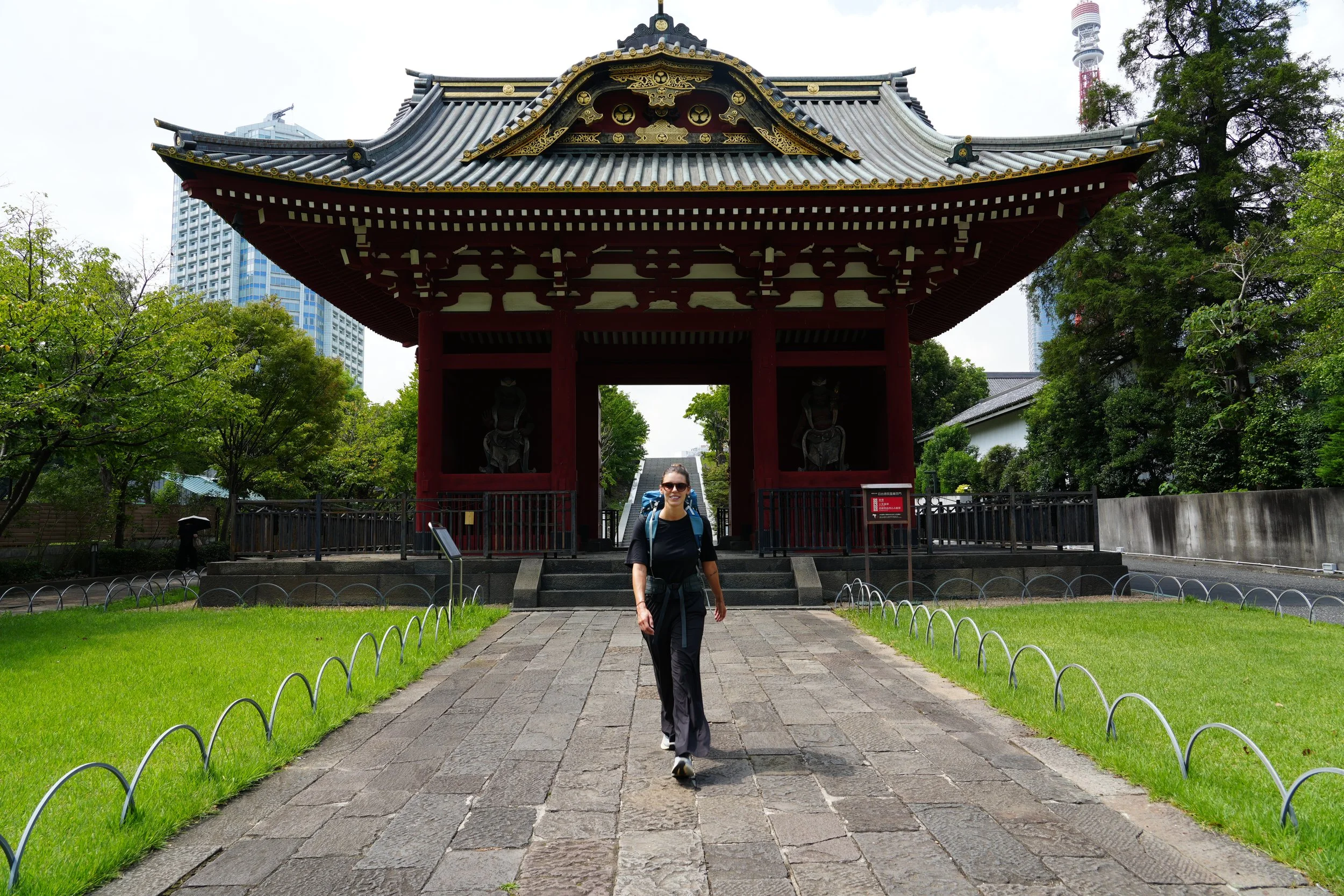 A woman in hiking gear walking on a stone pathway in front of a traditional Japanese gate with a layered, curved tiled roof, surrounded by green trees, with modern high-rise buildings and a tower visible in the background.