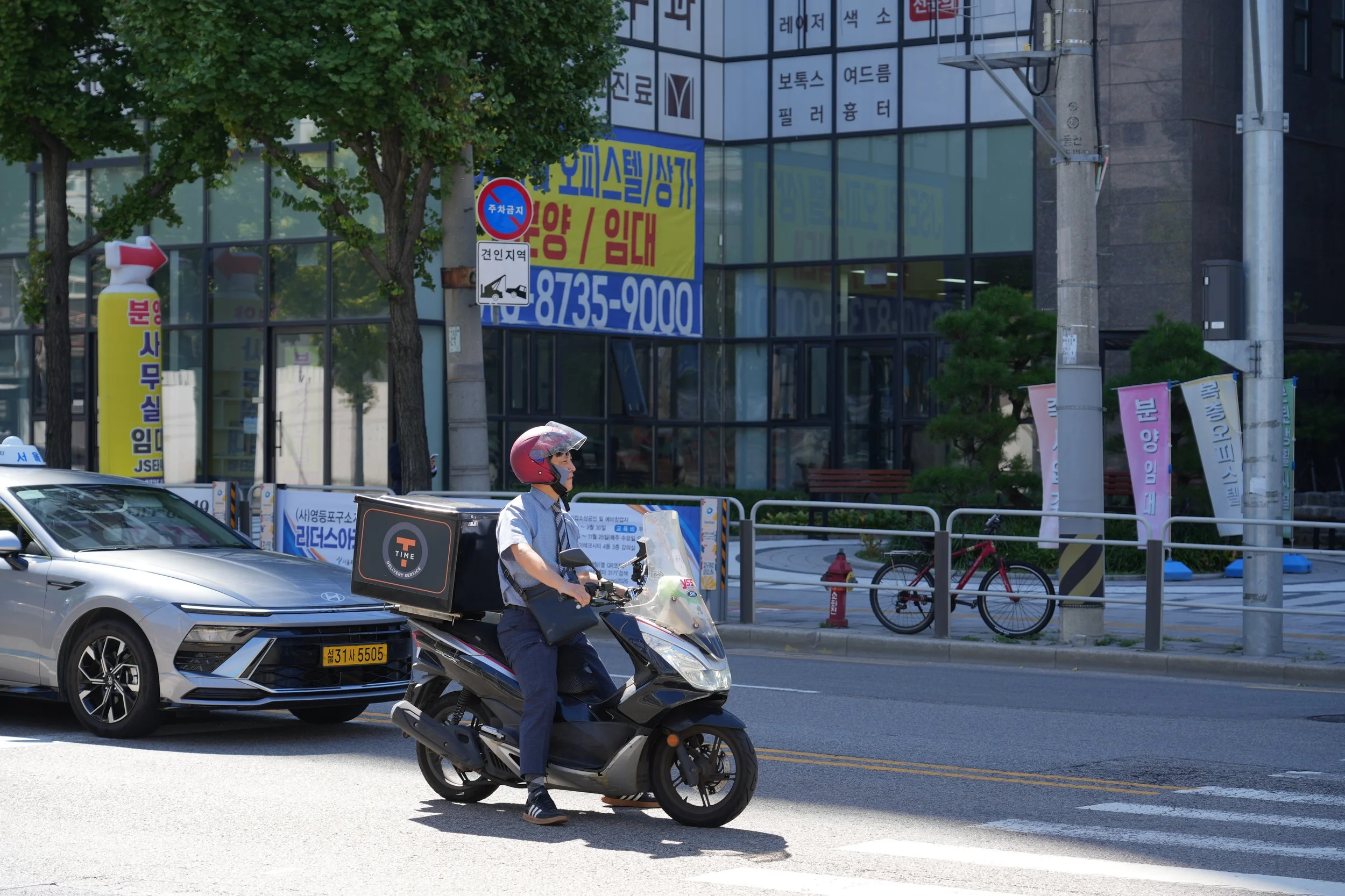 A person riding a scooter on a city street during the daytime, with a silver car parked nearby, trees, traffic signs, and a modern glass building in the background.