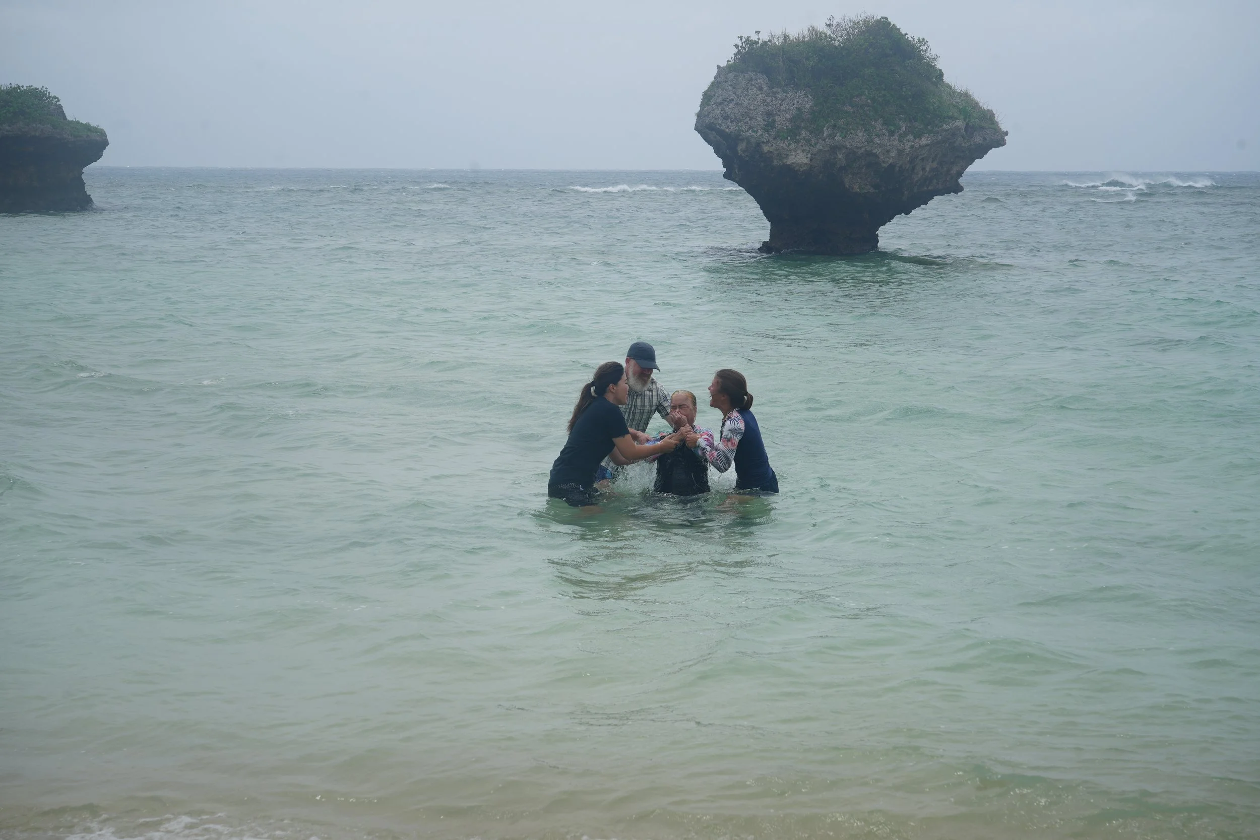 A group of five people standing in shallow water at the beach, holding hands and forming a circle. In the background are two large rock formations in the ocean, with a cloudy sky overhead.