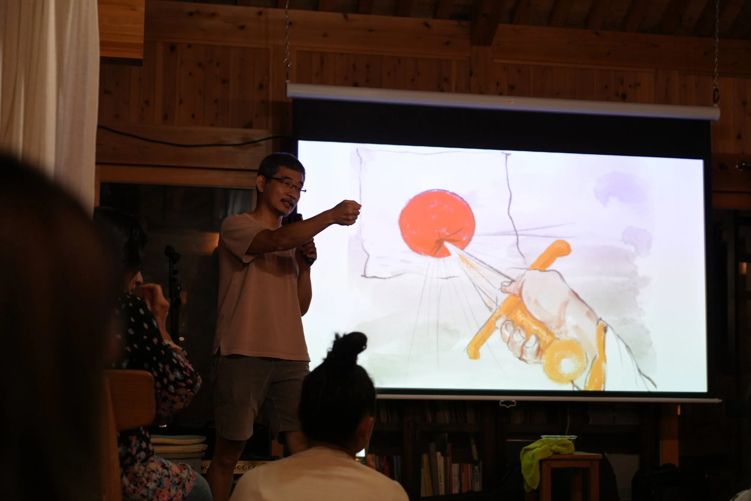 A man giving a presentation in front of a large screen displaying a colorful, abstract drawing. The room has wooden walls and a ceiling.