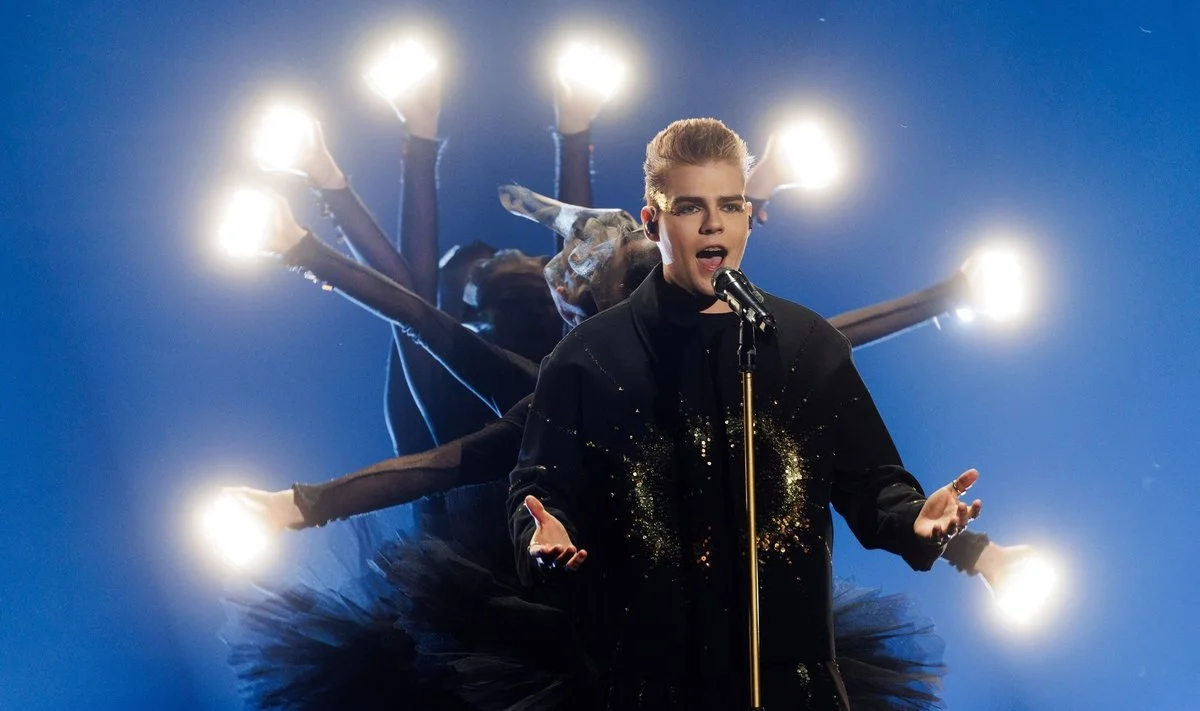 A performer singing into a microphone with multiple arms extending behind, each arm ending in a glowing light bulb, set against a dark blue background.