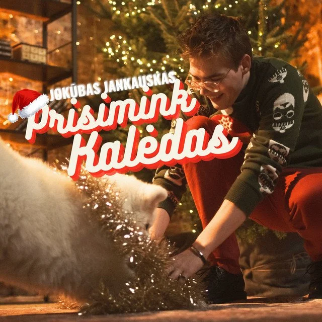 A young man smiling and playing with a dog in front of a decorated Christmas tree with presents and lights, with a Christmas-themed overlay text in Lithuanian