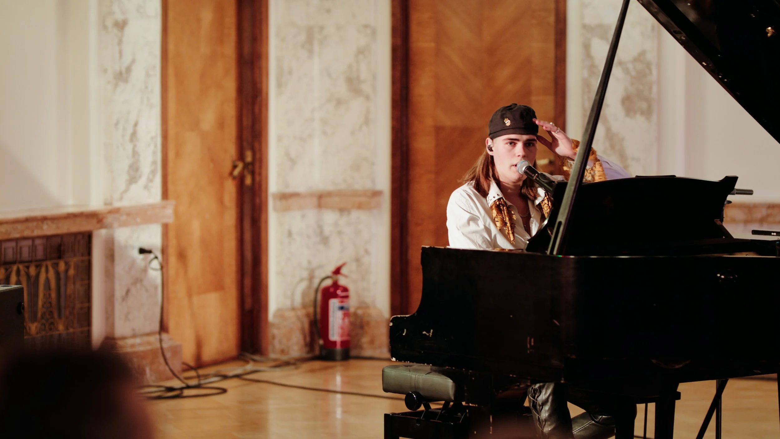 A young woman with long brown hair wearing a black cap and a patterned shirt, sitting at a black grand piano and singing into a microphone in a room with wood-paneled walls.