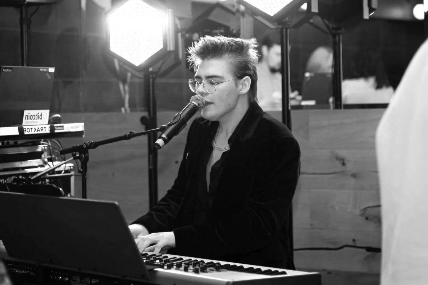 A young man with glasses and styled hair singing into a microphone while playing a keyboard in a dimly lit indoor setting.