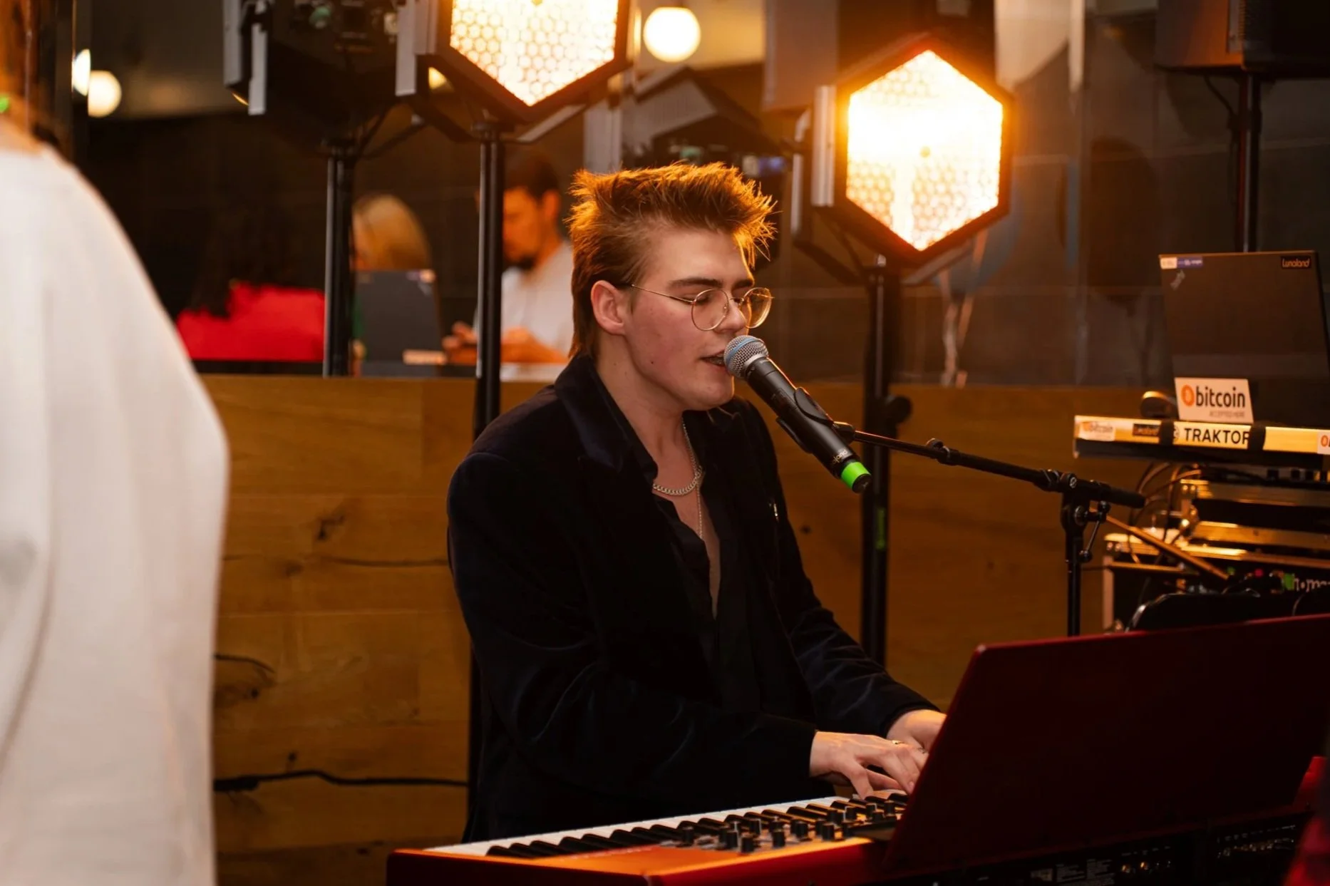 A young person with short, spiky hair, glasses, and wearing a black jacket, singing into a microphone while playing a keyboard in a warmly lit venue with wooden decor and musical equipment.