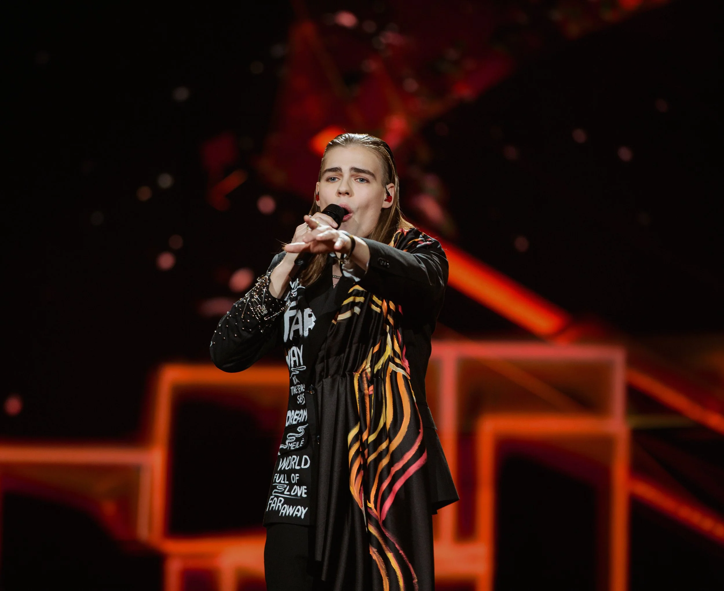 A female singer performing on stage, holding a microphone, wearing a black jacket with colorful stripes on the sleeves, standing against a dark background with red and orange stage lights.