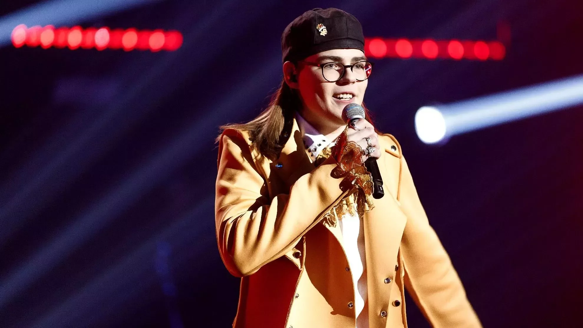 Person in glasses and a black cap holding a microphone speaking on stage, with bright stage lights in background.