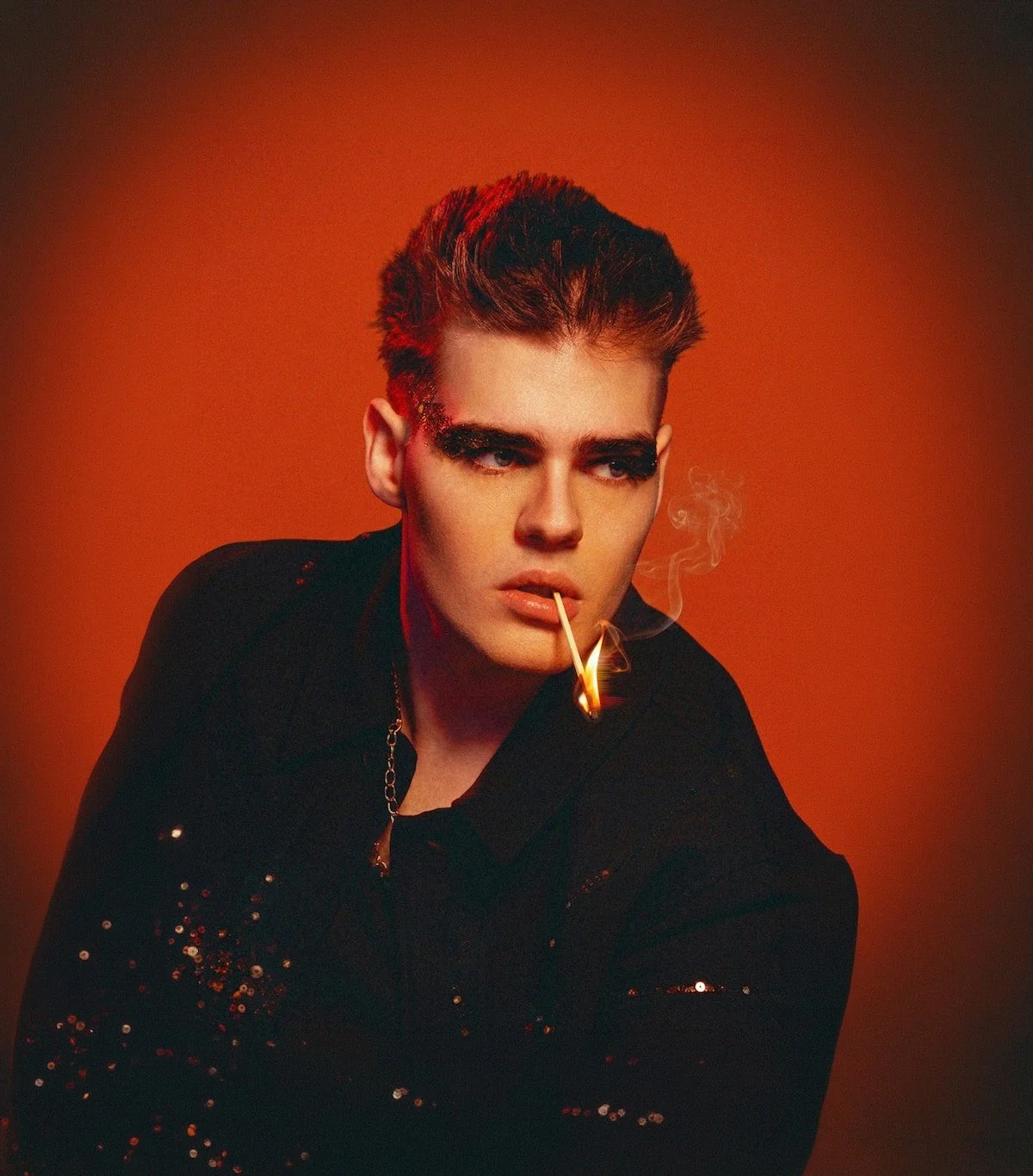 Young man with styled hair and bold makeup, wearing a black sequined jacket, has a lit match in his mouth, against a warm orange background.