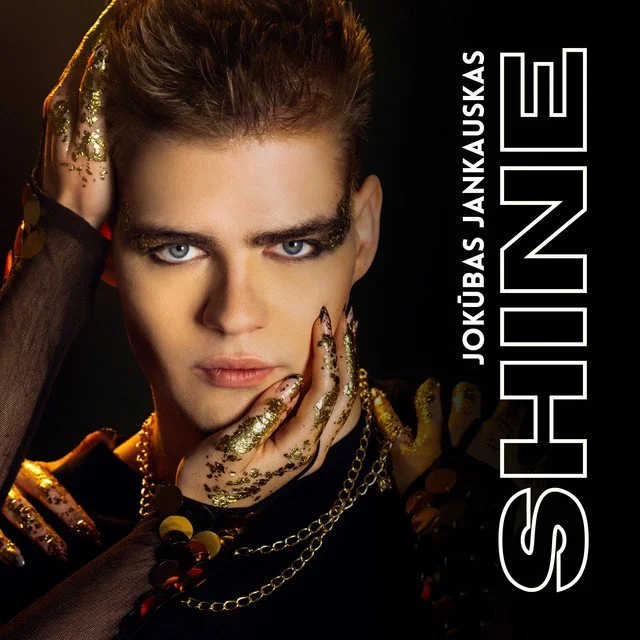 Close-up portrait of a young man with dramatic makeup and gold glitter on his hands and jewelry, with the name 'SHUNE' and 'JOKUBAS JANKAUSKAS' written vertically beside him.