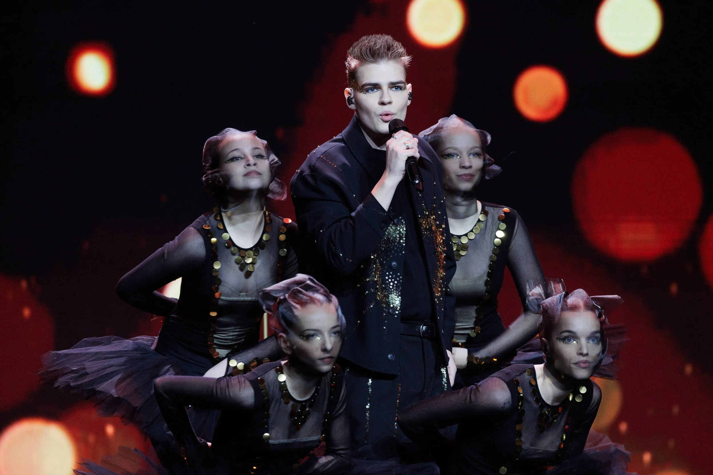 A male performer singing into a microphone on stage, surrounded by female dancers in black costumes with gold sequins and tulle skirts, with a dark background and red and orange stage lighting.