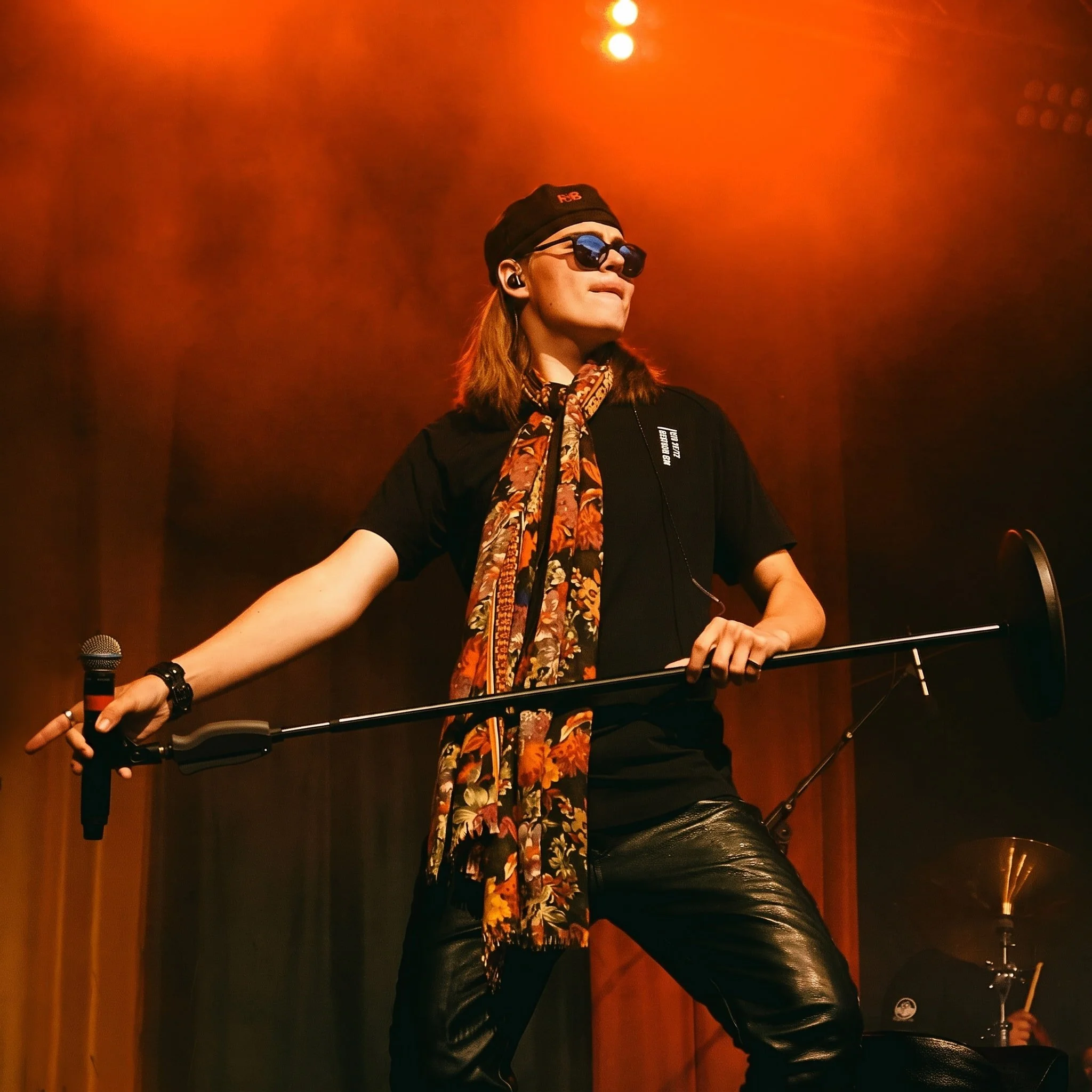Person wearing sunglasses and a black cap, with long hair, at a music event or nightclub with red lighting, holding a microphone in one hand and a stick in the other, standing next to a DJ setup.