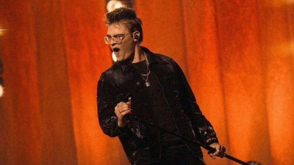 A young man performing on stage, singing into a microphone, with a passionate expression, wearing glasses, a black jacket, and a necklace, in front of orange stage curtains.