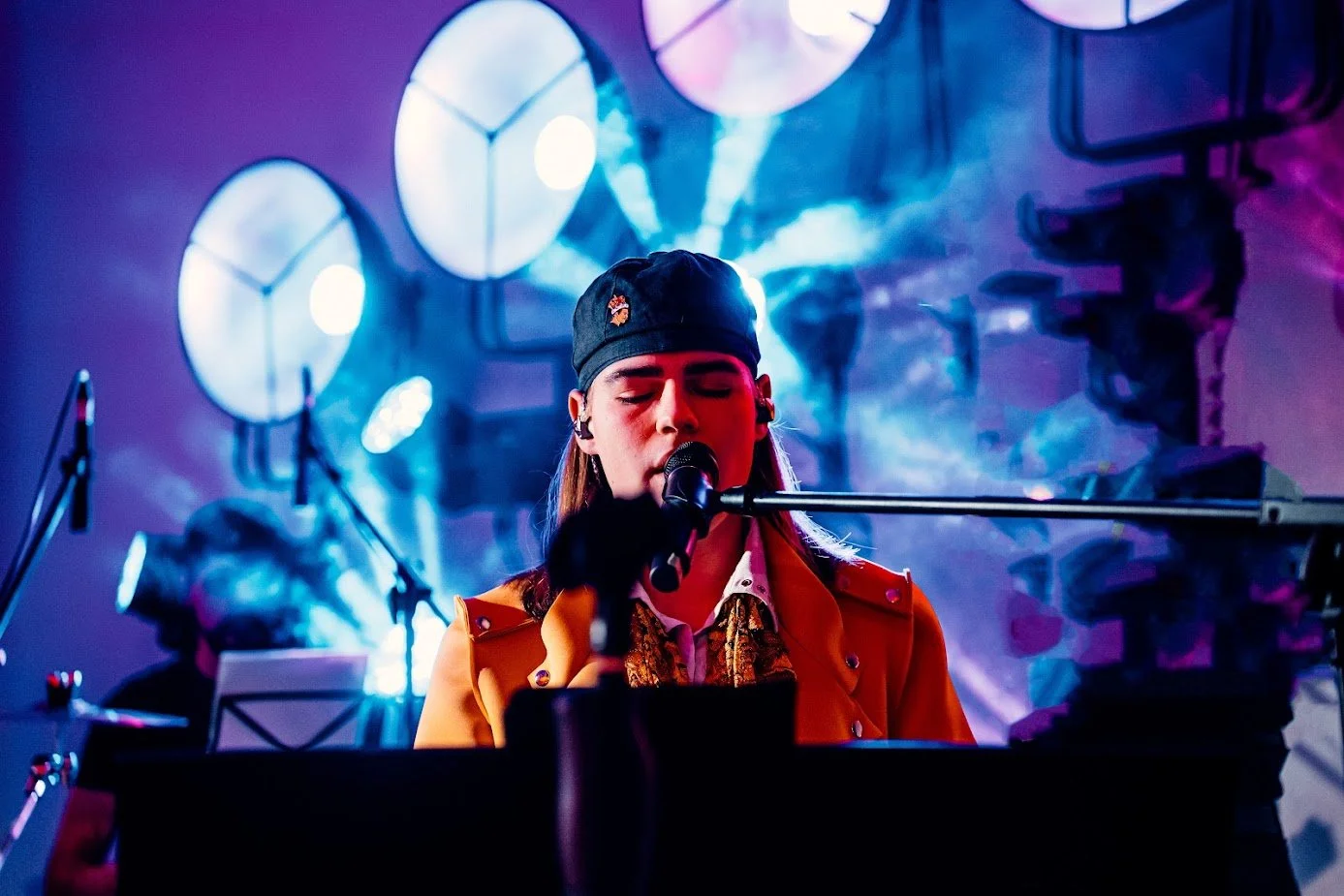 A woman singing into a microphone while playing the piano on stage with blue and purple lighting and circular backdrop lights.