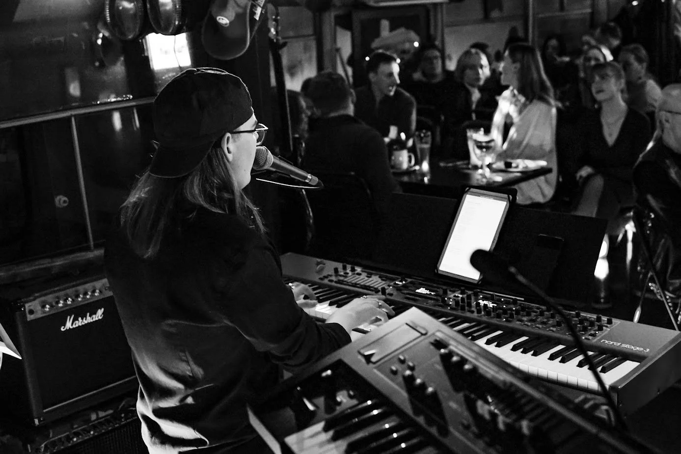 A woman in a black baseball cap and glasses playing a keyboard and singing into a microphone at a performance in a dimly lit venue, with an audience in the background.