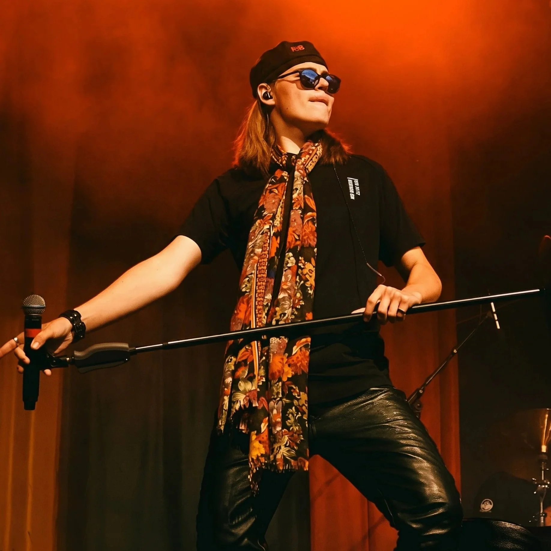 A man with long hair wearing sunglasses, a black beanie, and a colorful scarf, standing on a stage with red lighting, with a microphone.